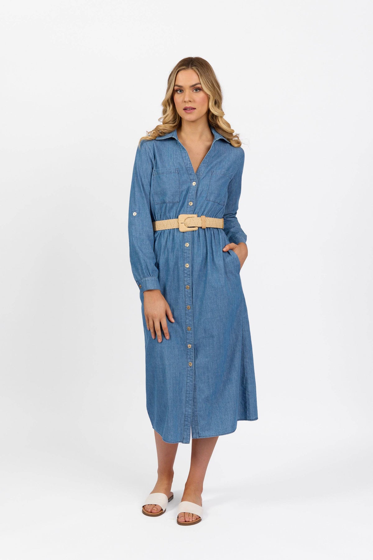A woman with blonde, wavy hair stands against a white background wearing the Vassalli Long Sleeve Dress - Blue Wash, styled with a beige belt and white slide sandals. She has one hand in her pocket.