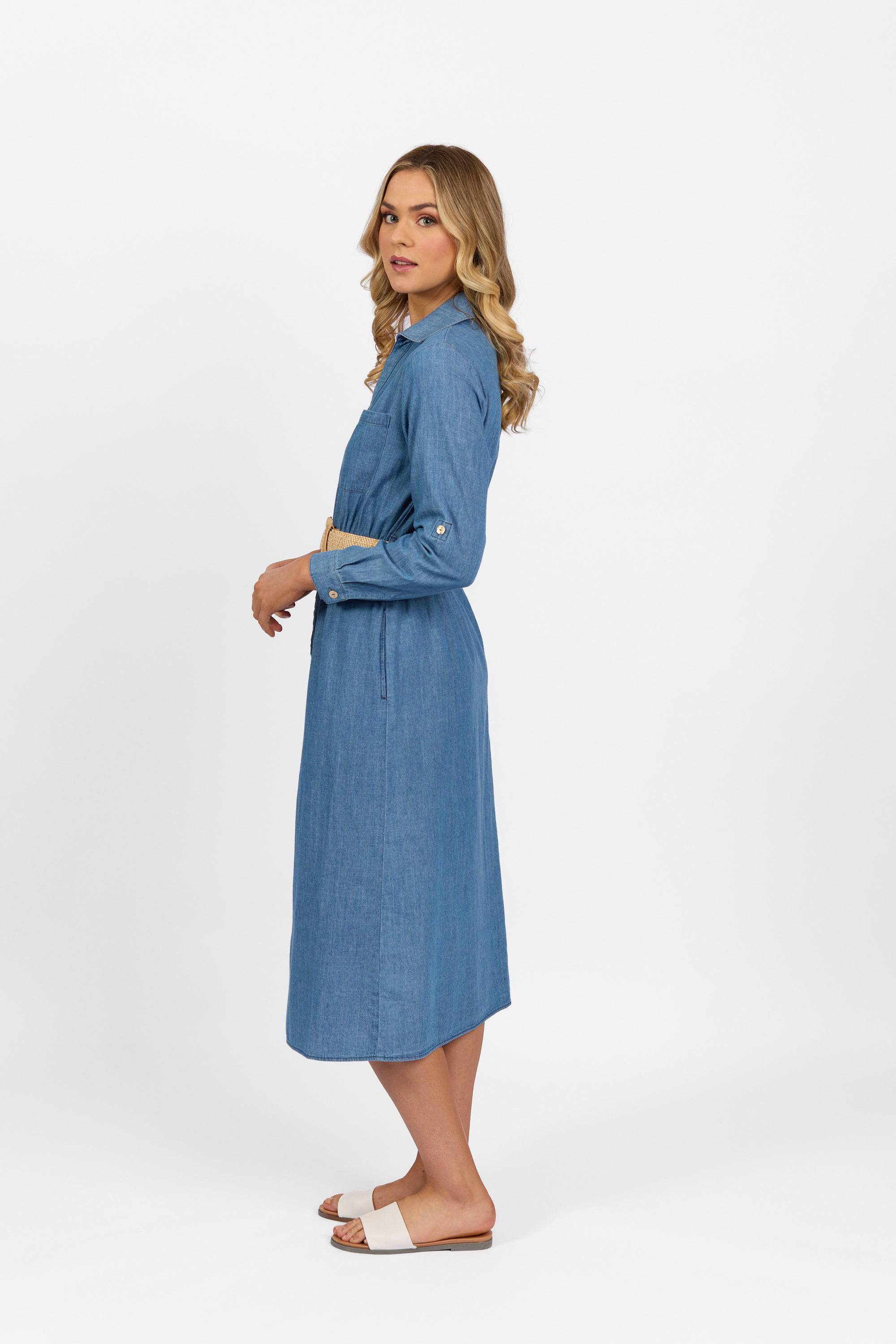 A woman with long wavy hair stands sideways in the Vassalli Long Sleeve Dress - Blue Wash and light-colored slide sandals, against a plain white background.