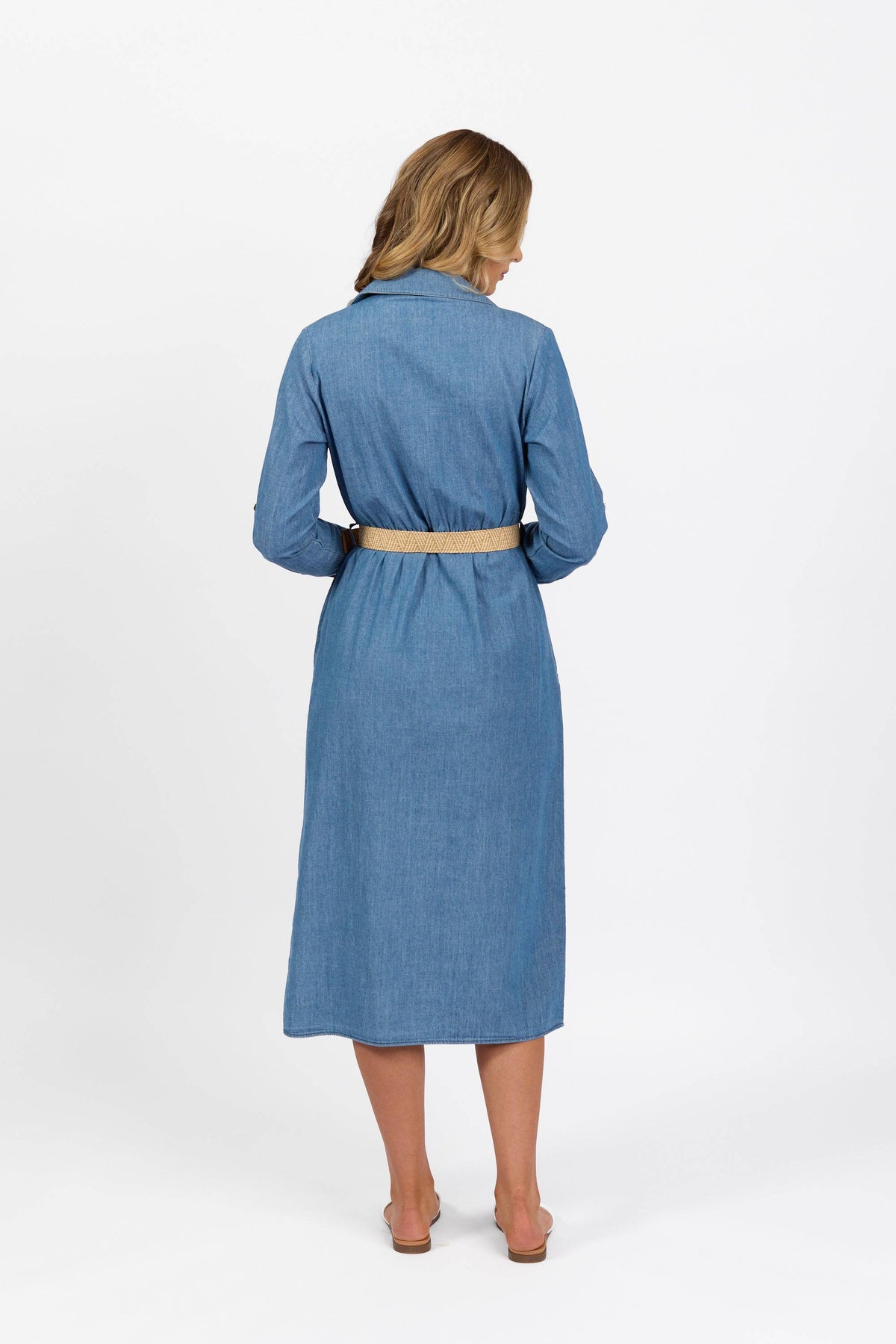A woman with shoulder-length blonde hair stands barefoot on a white background, wearing the Vassalli Long Sleeve Dress - Blue Wash with a beige belt.