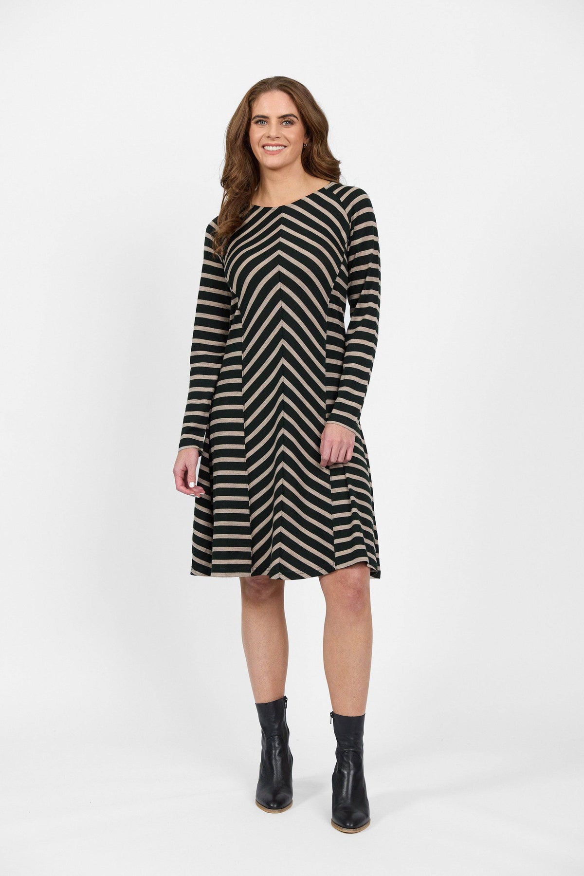 A woman with long brown hair smiles against a white background wearing the Vassalli Swing Merino Dress in Forest/Twig Stripe, paired with black ankle boots.
