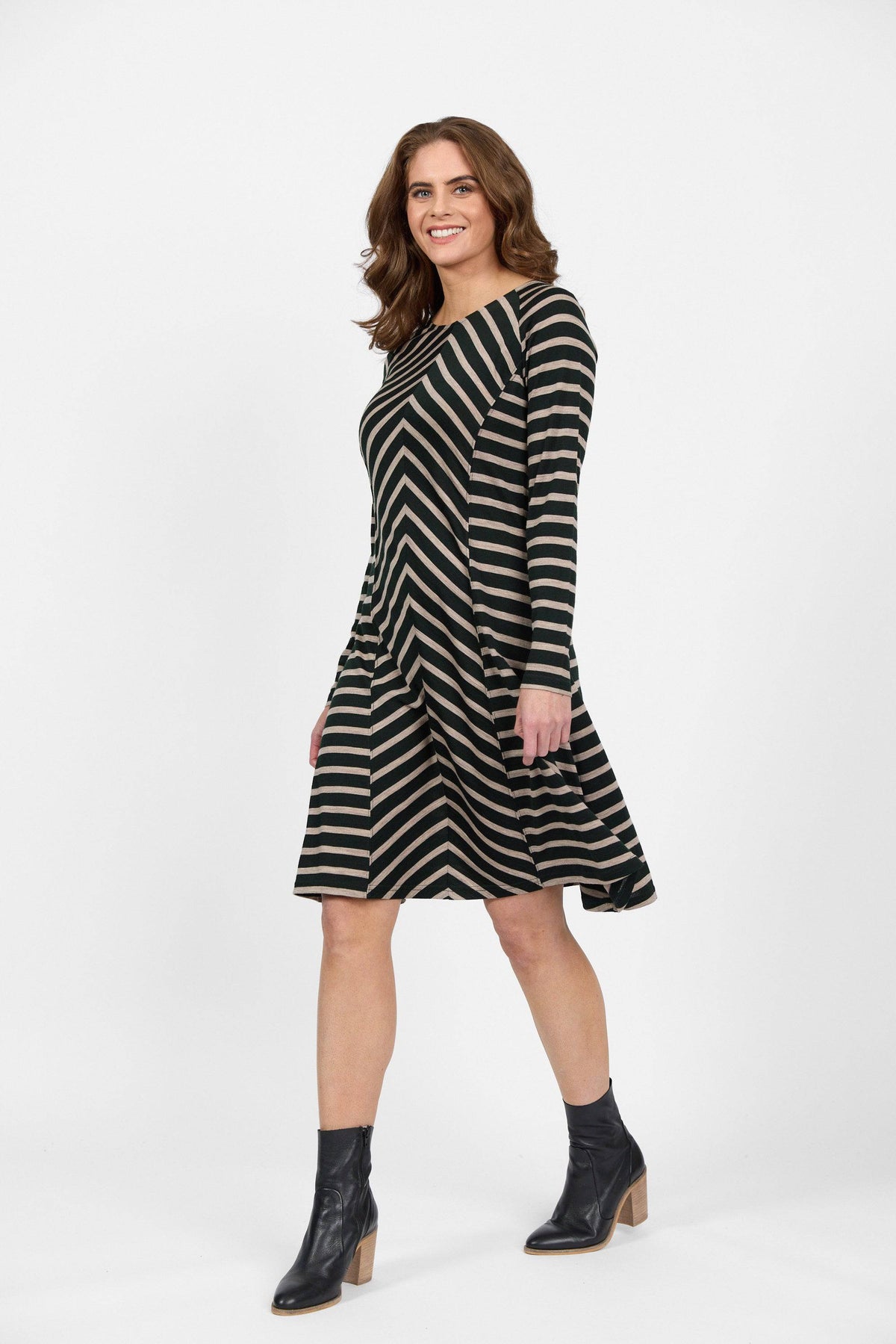 A woman smiles while walking in the Vassalli Swing Merino Dress - Forest/Twig Stripe, featuring diagonal green and beige stripes, paired with black ankle boots against a plain white background.