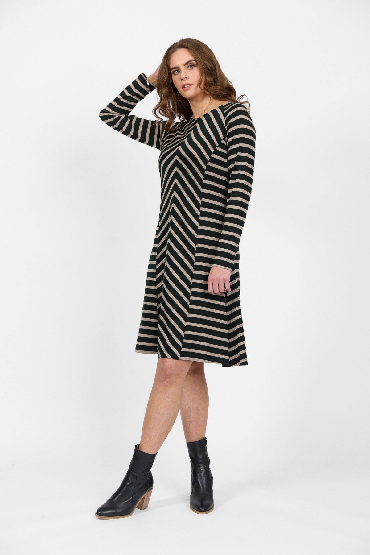 Woman stands against a white background, wearing the Vassalli Swing Merino Dress in Forest/Twig Stripe with black ankle boots. She raises one hand to her head and looks at the camera.