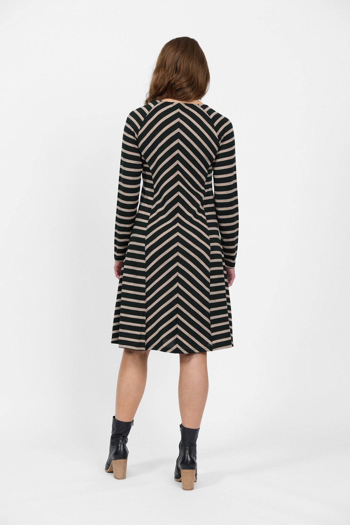 A woman with mid-length brown hair wears the Vassalli Swing Merino Dress in Forest/Twig Stripe, paired with black chunky-heeled ankle boots, standing against a plain white background, facing away from the camera.