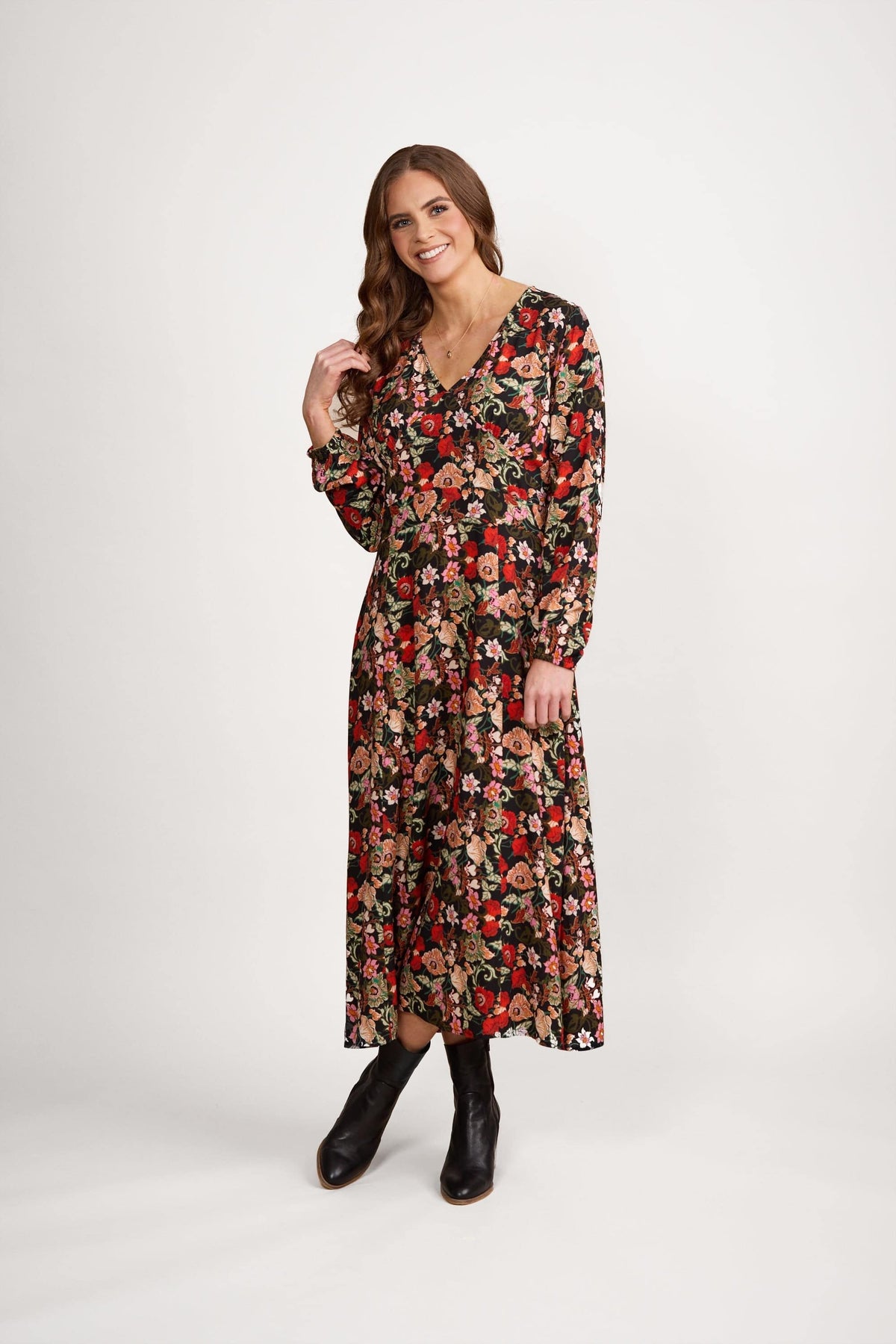 A woman with long brown hair models the Vassalli V-Neck Dress - Flamenco, featuring long sleeves and an A-line skirt with red, pink, and beige floral print on black. She pairs it with black ankle boots and smiles against a plain white background.