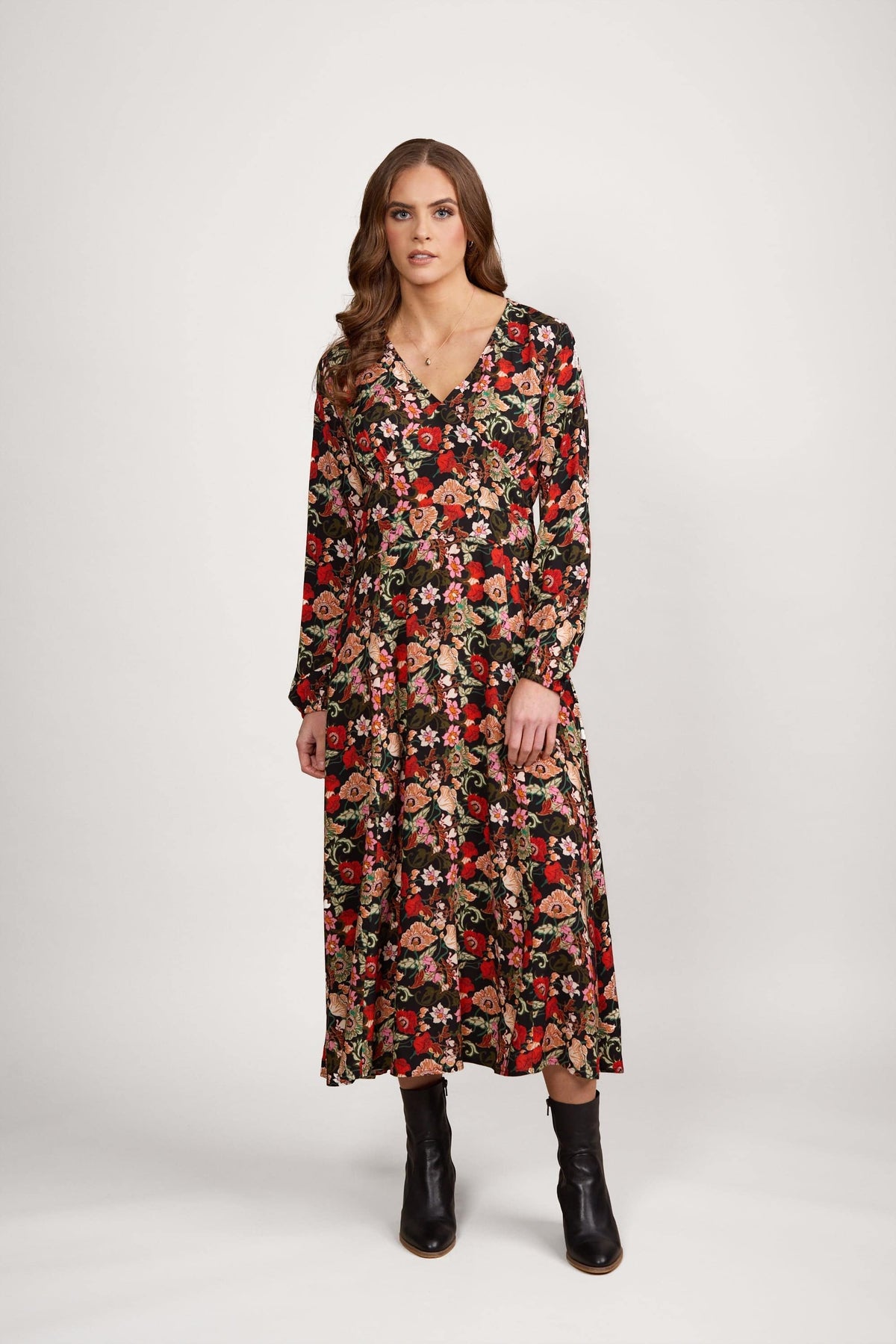 A woman with wavy brown hair wears the Vassalli V-Neck Dress - Flamenco, featuring long sleeves and an A-line skirt in red, pink, and green florals. She pairs it with black ankle boots against a plain white background.