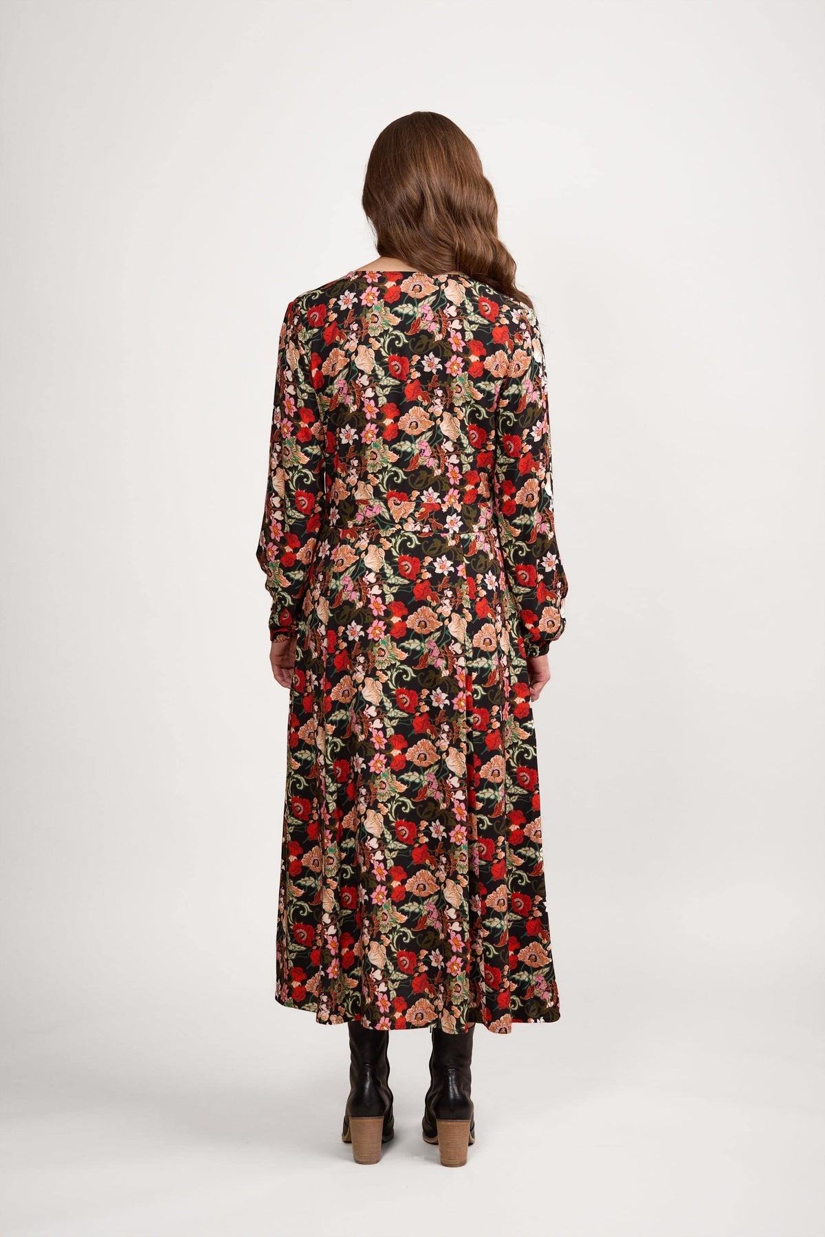 A woman with long brown hair, seen from behind, wears the Vassalli V-Neck Dress - Flamenco with a floral print and black boots, standing against a plain white background.