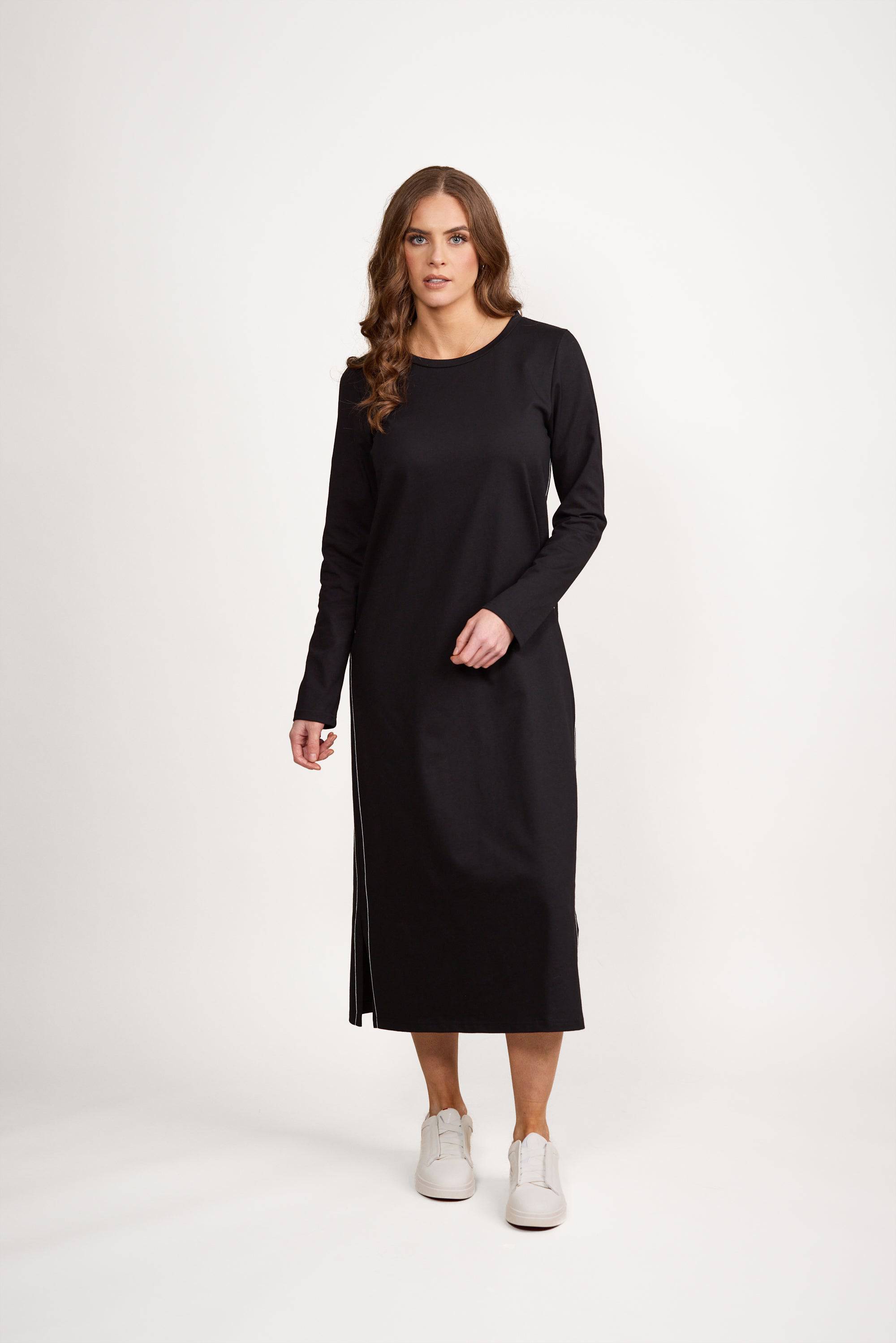 A woman stands facing forward, wearing a relaxed, loose-fit Vassalli Knit Dress - Black/White in ankle length, paired with white sneakers, against a plain white background.