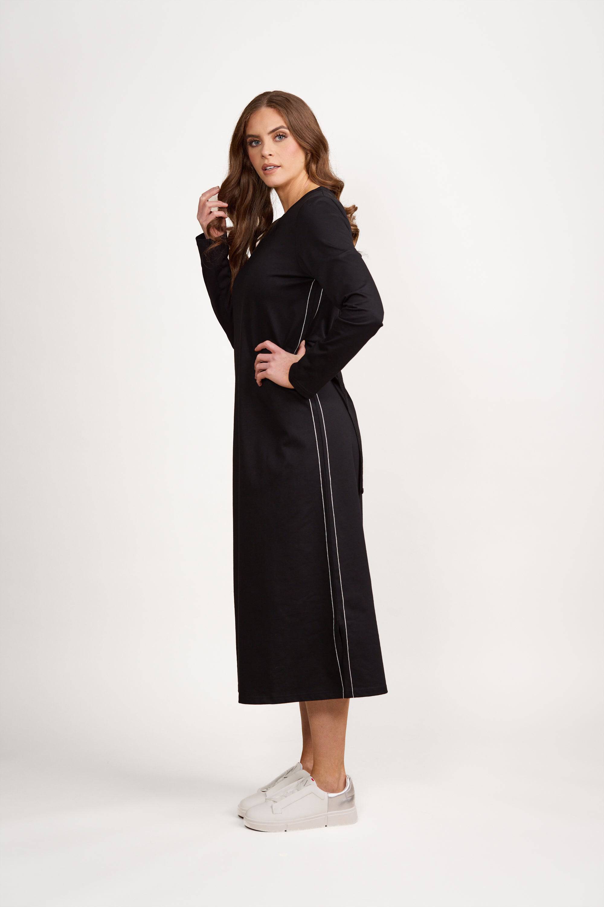 A woman stands sideways against a plain white background, wearing the Vassalli Knit Dress - Black/White by Vassalli. The soft knit dress features thin white side stripes and a relaxed, loose fit. She pairs it with white sneakers and looks at the camera.