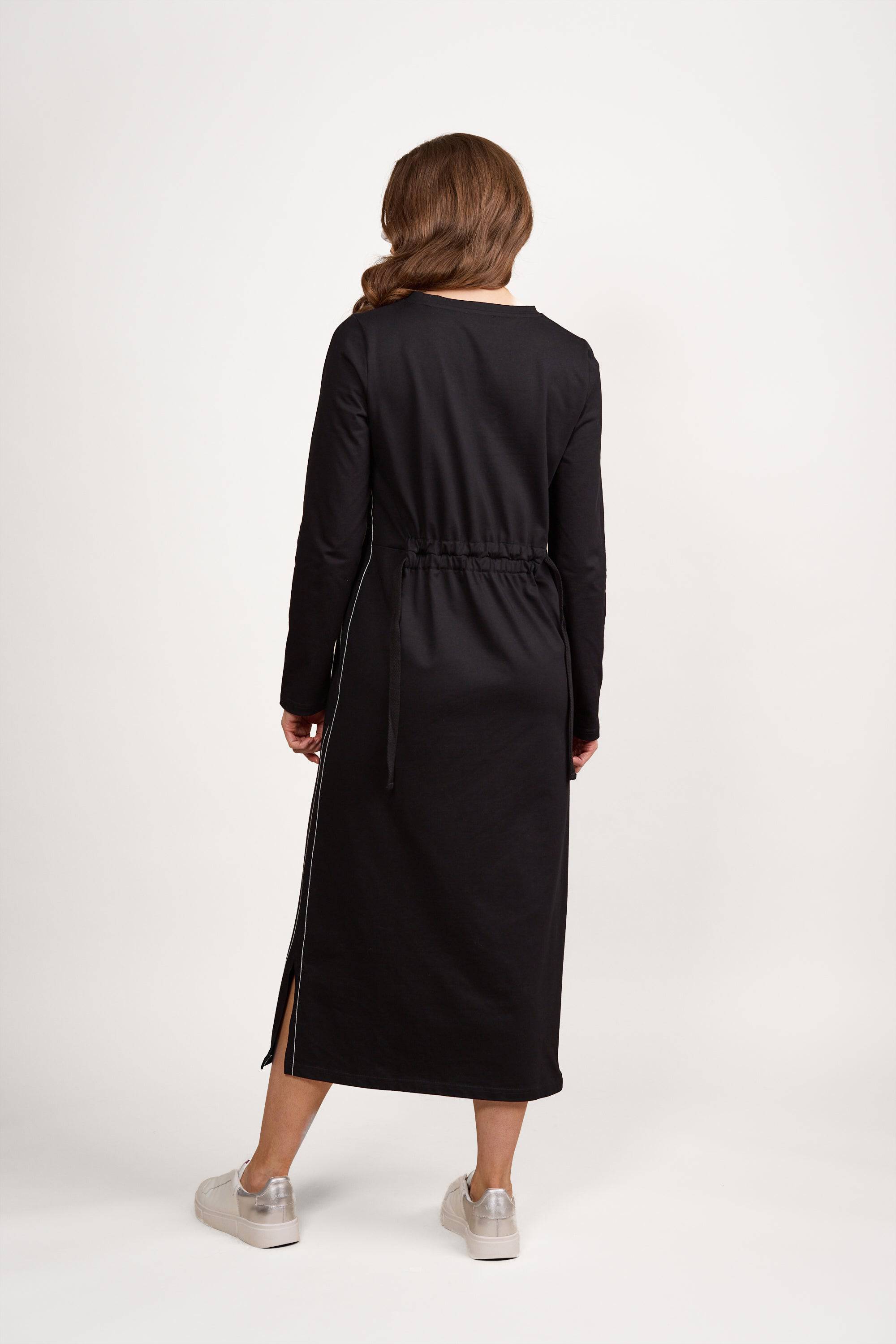 A person with long brown hair stands facing away, wearing the Vassalli Knit Dress - Black/White in a relaxed loose fit with a drawstring waist and side slit, crafted from soft knit fabric, paired with silver sneakers, against a plain white background.