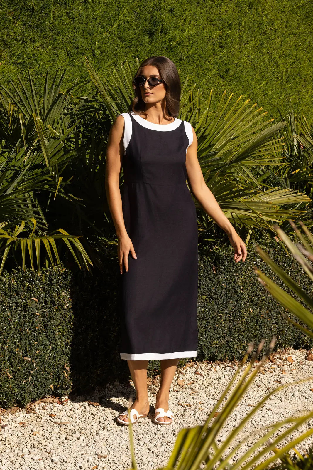 A woman stands on gravel in front of lush green bushes and palm leaves, sunlight casting shadows, wearing the Vassalli Oak Dress in Ink/White—a sleeveless summer dress—paired with white sandals and sunglasses.