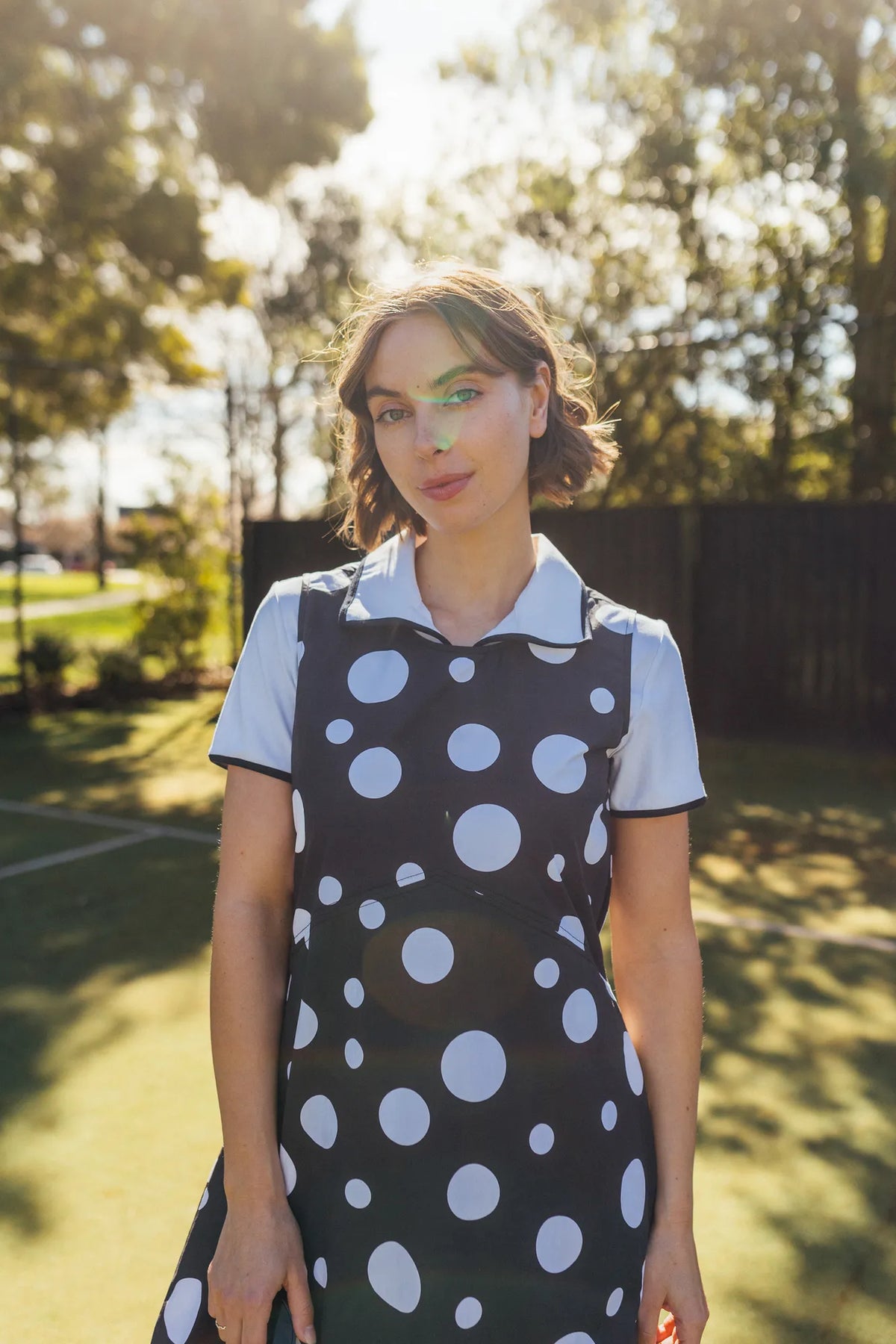 A person stands outside in sunlight, wearing the Vassalli Blake Dress - Spot, a sleeveless cotton dress with a black and white spot print, layered over a white collared shirt. Green trees and a blurred fence are visible in the background.