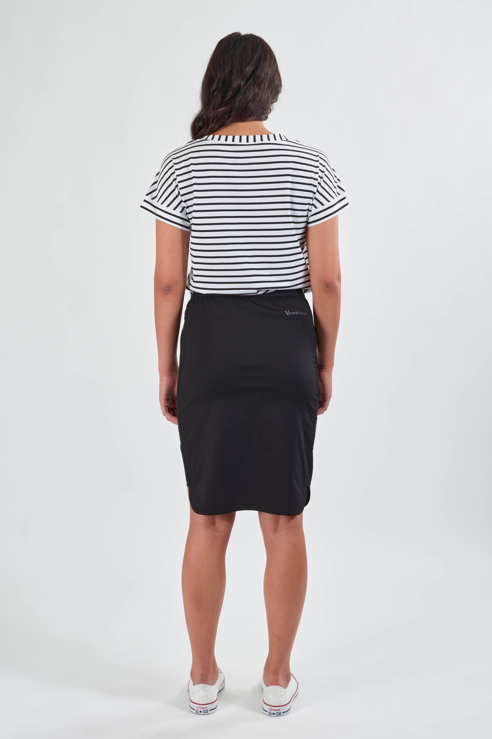 A woman faces away, wearing the Vassalli Poplin Comfort Skirt in black, paired with a short-sleeved white top with black stripes and white slip-on shoes, set against a plain light background.
