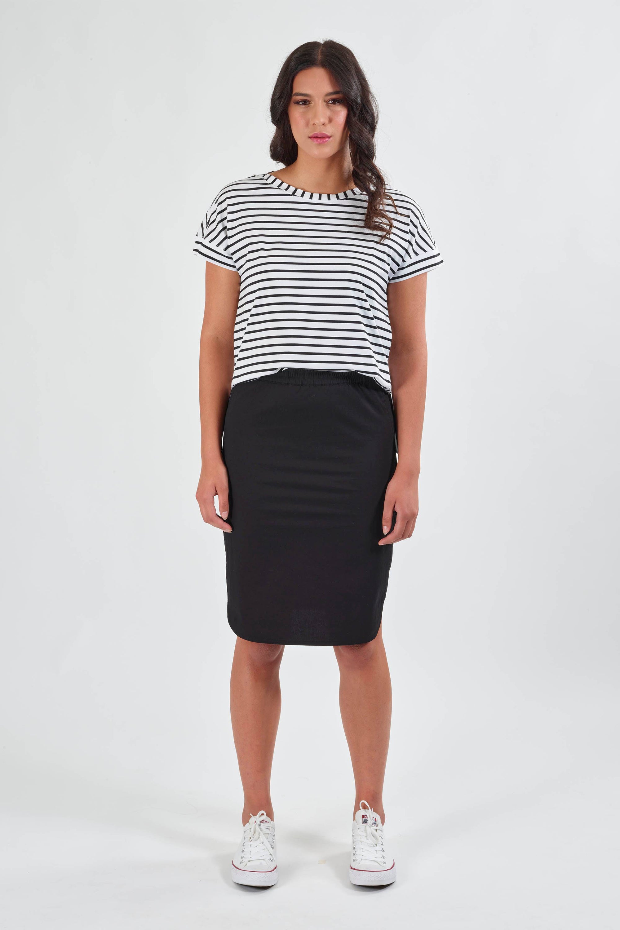 A woman faces forward against a plain white background, wearing the Vassalli Poplin Comfort Skirt in black, a black-and-white striped short-sleeve shirt, and white sneakers.