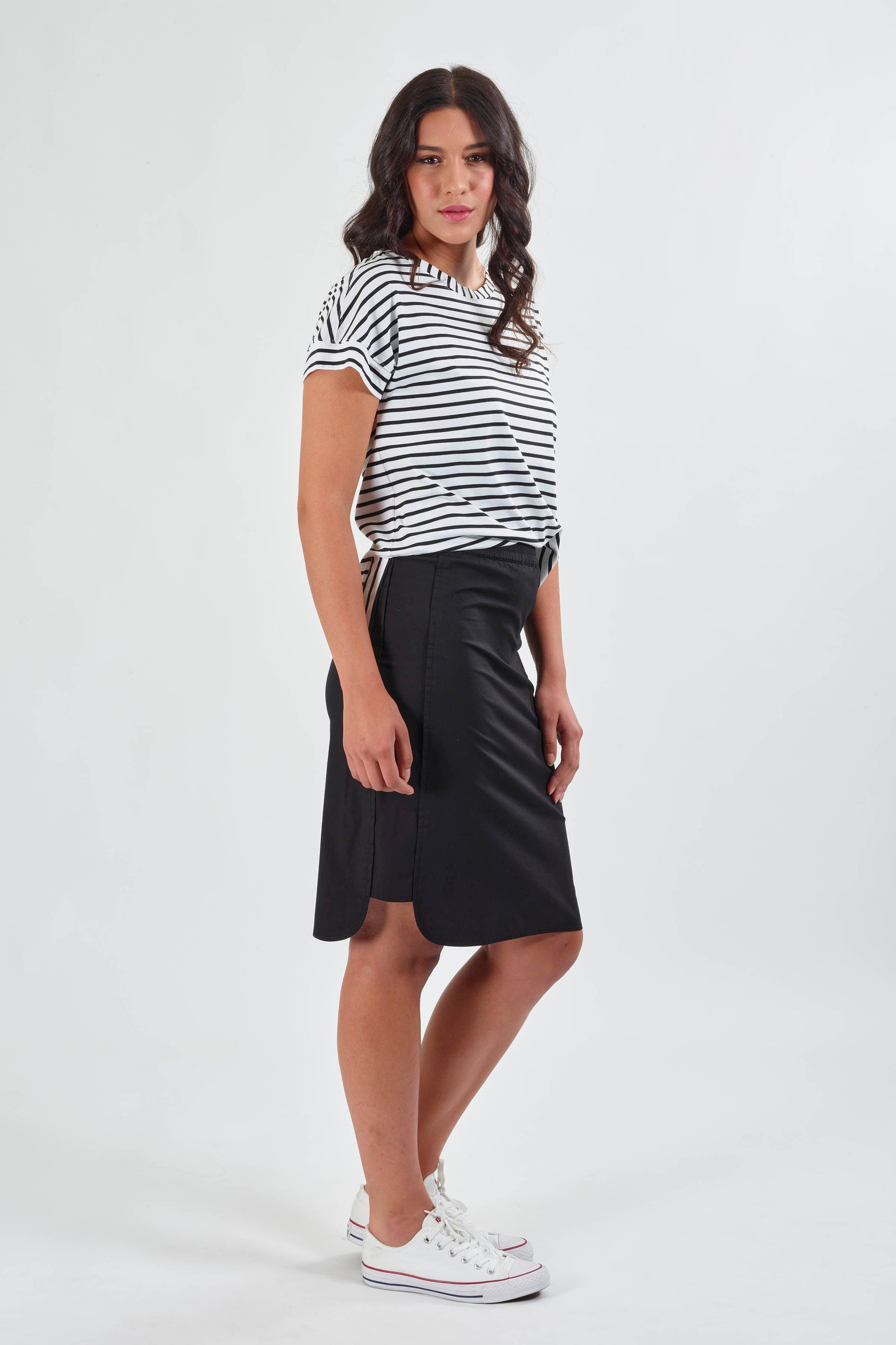 A woman with long wavy hair and a neutral expression models the Vassalli Poplin Comfort Skirt in black, paired with a white and black striped short-sleeve top and white sneakers against a plain white background.