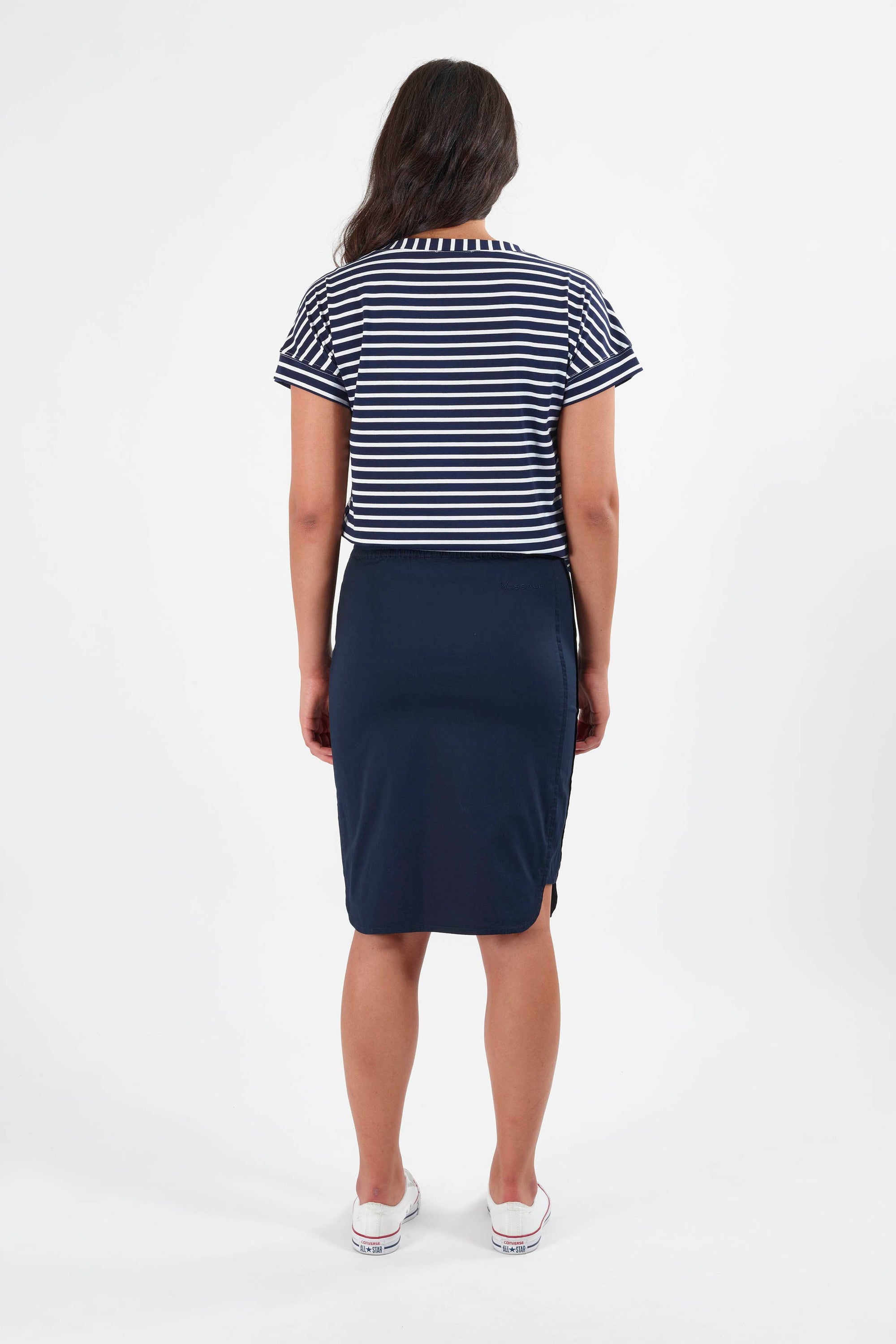 A woman stands facing away against a white background, wearing a navy blue and white striped short-sleeve top, Vassalli’s Poplin Comfort Skirt - Navy, and white sneakers.