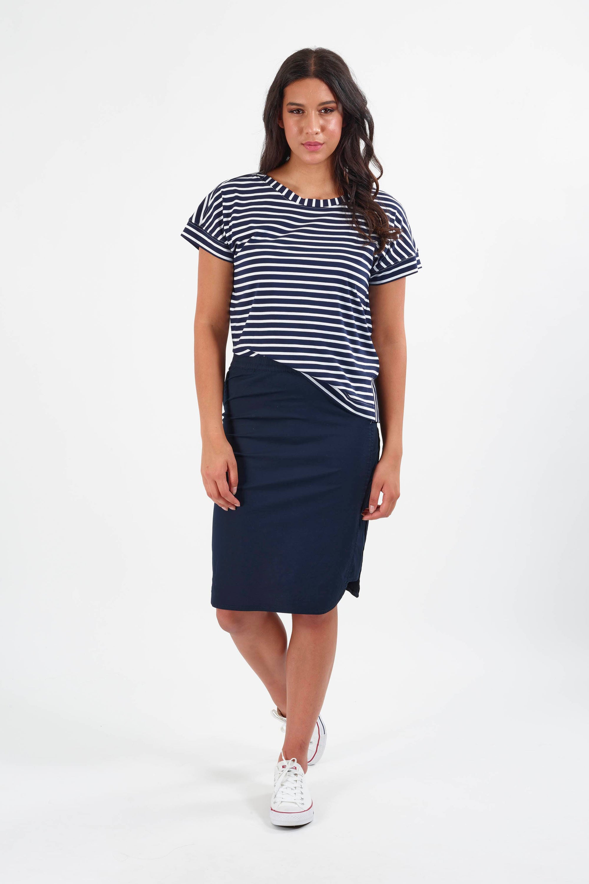 A woman with long dark hair and a relaxed expression wears the Vassalli Poplin Comfort Skirt in navy, paired with a navy and white striped t-shirt and white sneakers, standing against a white background.
