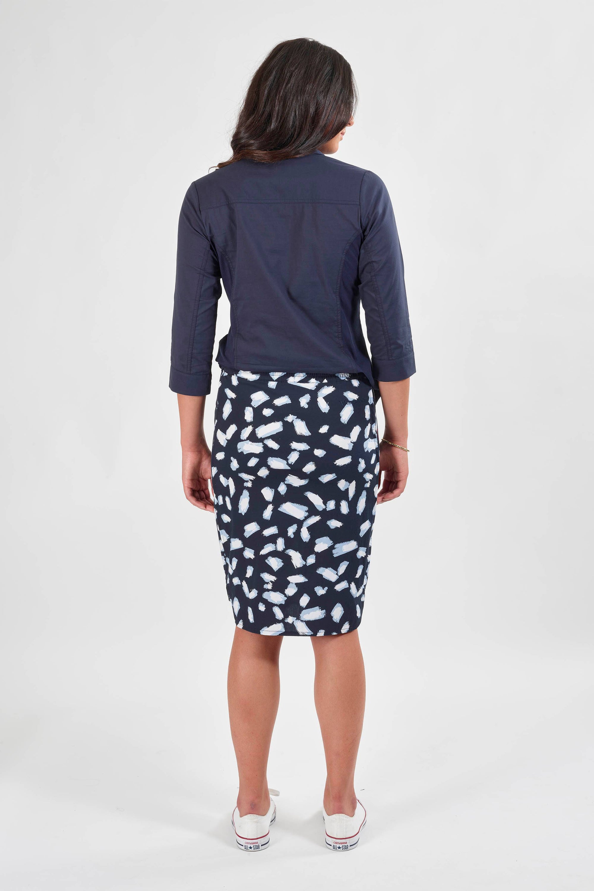A woman faces away from the camera wearing a navy top, white sneakers, and the Vassalli Poplin Comfort Skirt - Shore, featuring a knee-length white and navy abstract pattern, set against a plain white background.