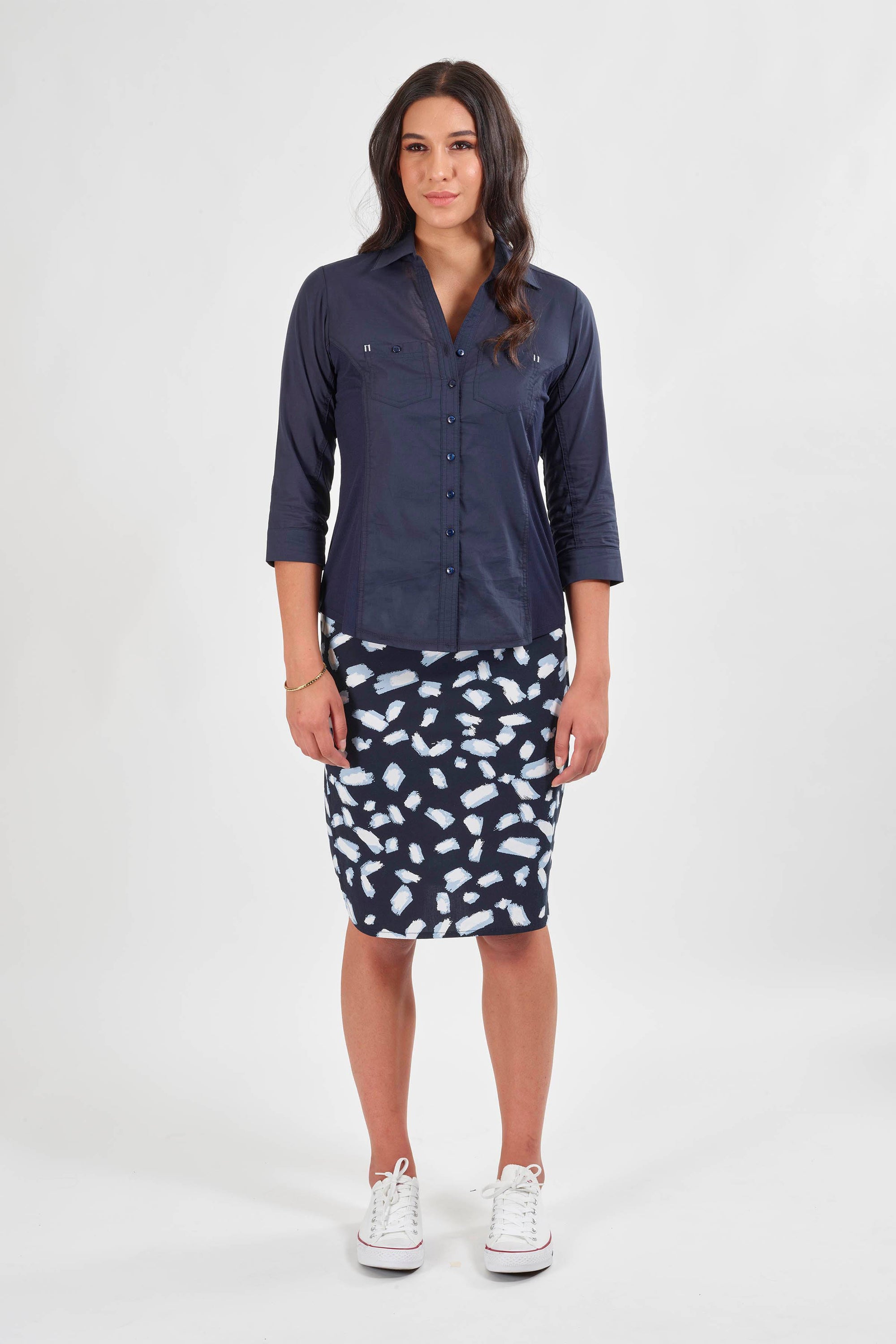 A woman with long, wavy dark hair stands against a plain white background, wearing a dark blue button-up shirt, white sneakers, and the Vassalli Poplin Comfort Skirt in Shore—a navy and white knee-length skirt.