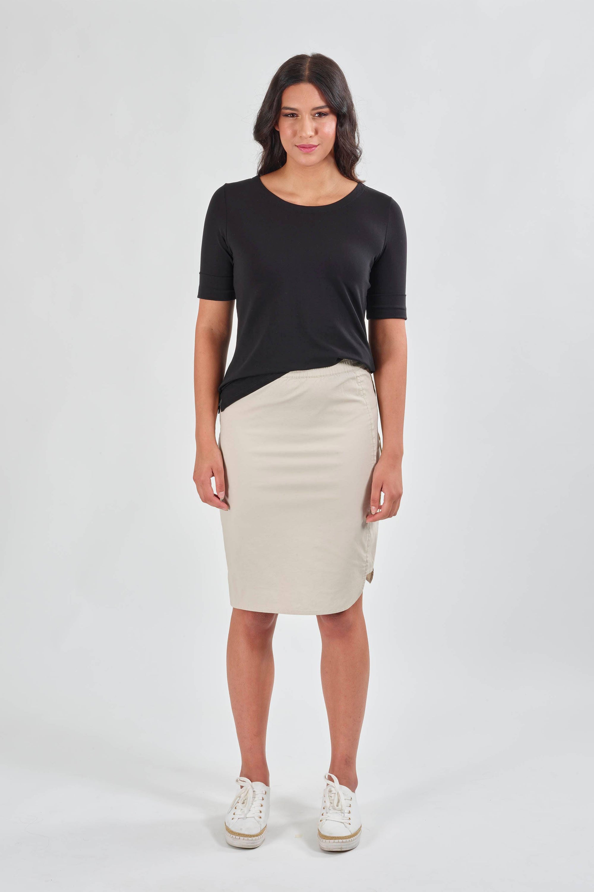 A woman faces forward in a black short-sleeve top, Vassalli Poplin Comfort Skirt - Sand, and white sneakers against a plain light backdrop.