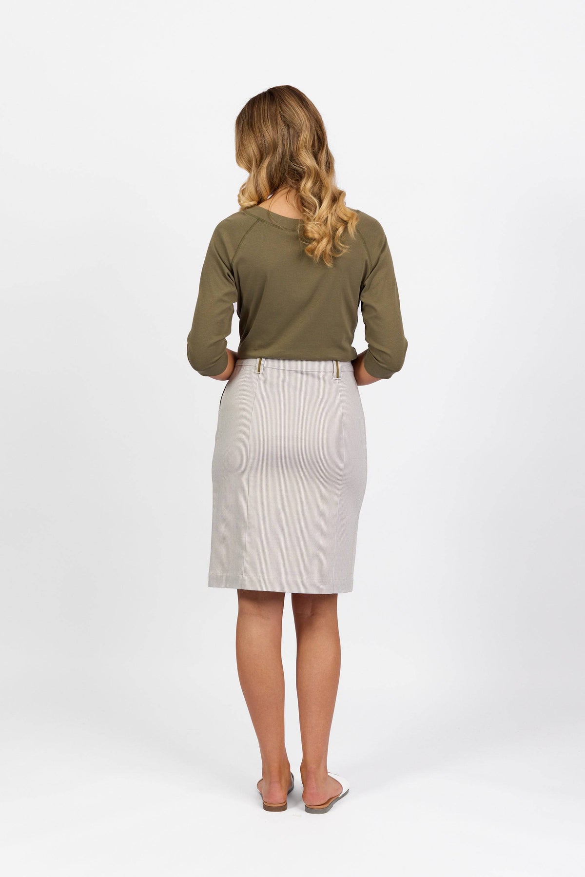 A woman with wavy blonde hair stands facing away from the camera against a plain white background, wearing an olive green long-sleeve top, sandals, and the Vassalli Orion Skirt in Khaki Stripe.
