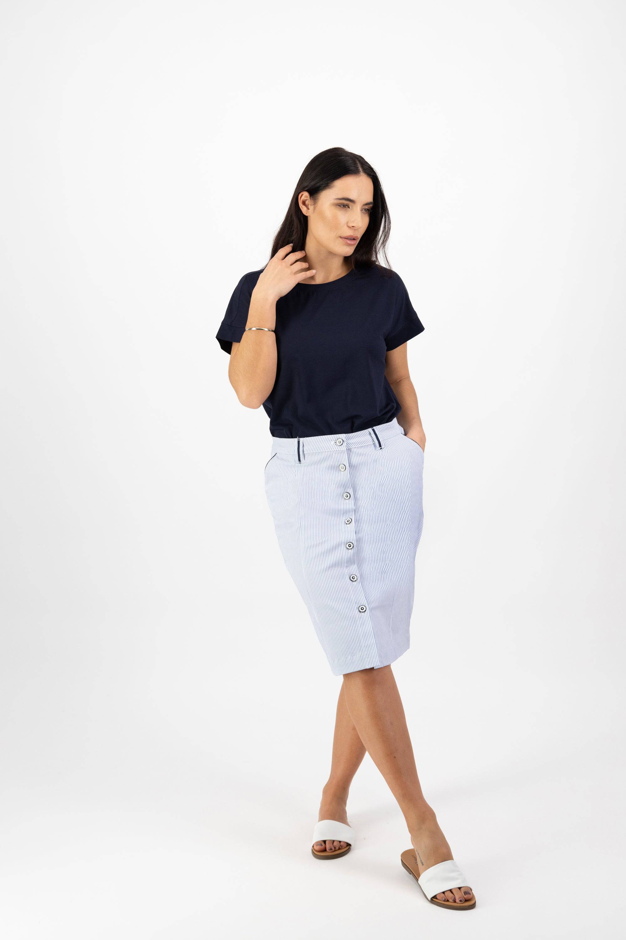 A woman with long dark hair stands against a white background, wearing a navy blue t-shirt, Vassalli Orion Skirt - Blue Stripe with pockets, white sandals, and a bracelet. One hand touches her hair while the other rests in her pocket.
