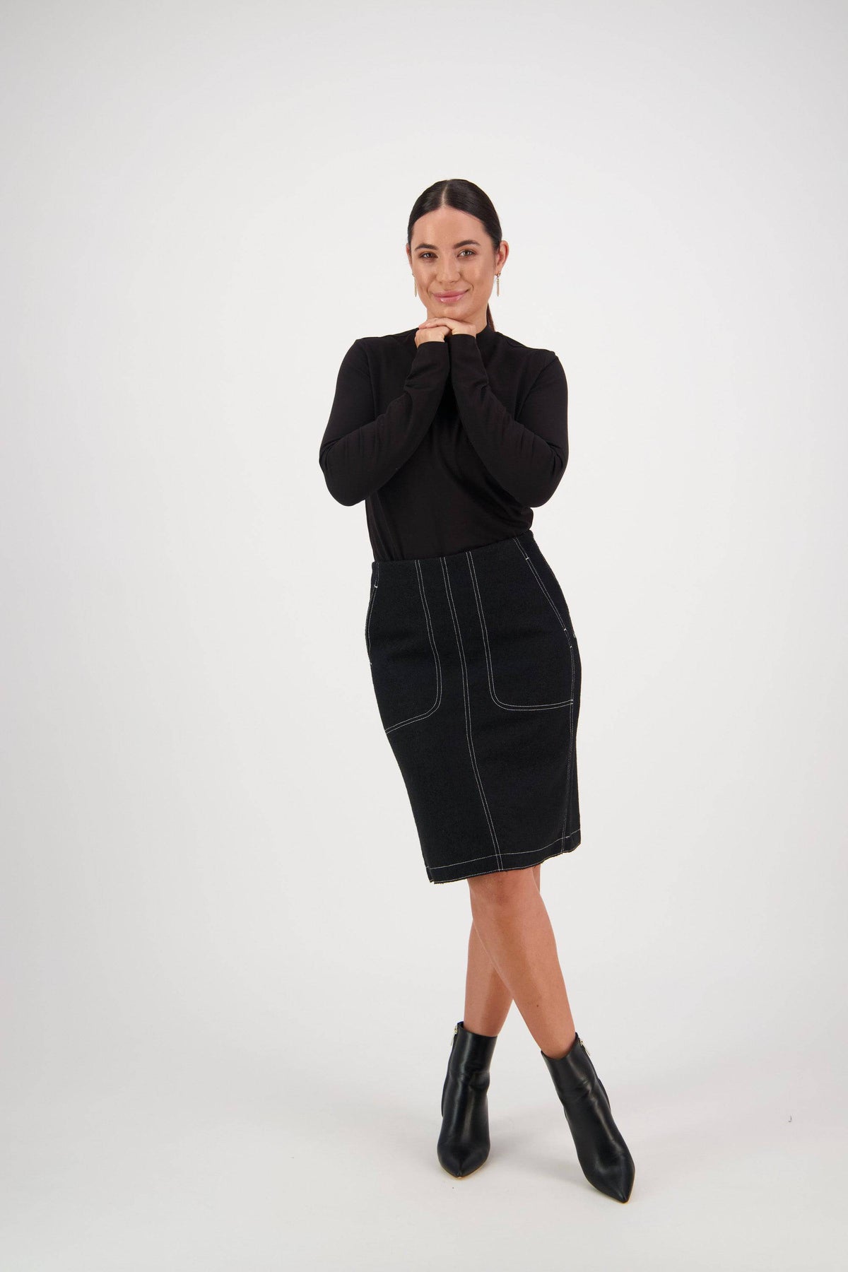 A person wearing a black long-sleeve top, the Vassalli Knee Length Skirt - Black with white stitching and front patch pockets, and black ankle boots stands with hands under chin, smiling against a plain white background.