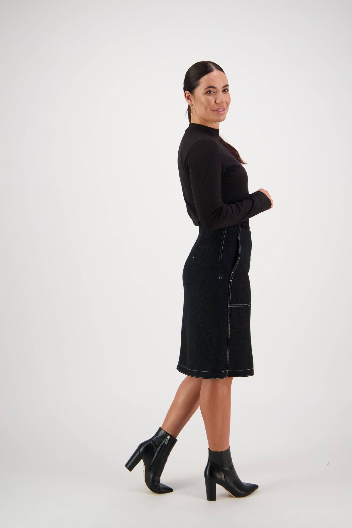 A woman stands sideways against a plain background, smiling with her hair in a ponytail. She wears a long-sleeve black top paired with the Vassalli Knee Length Skirt – Black, featuring visible seams and front patch pockets, styled with black heeled ankle boots.