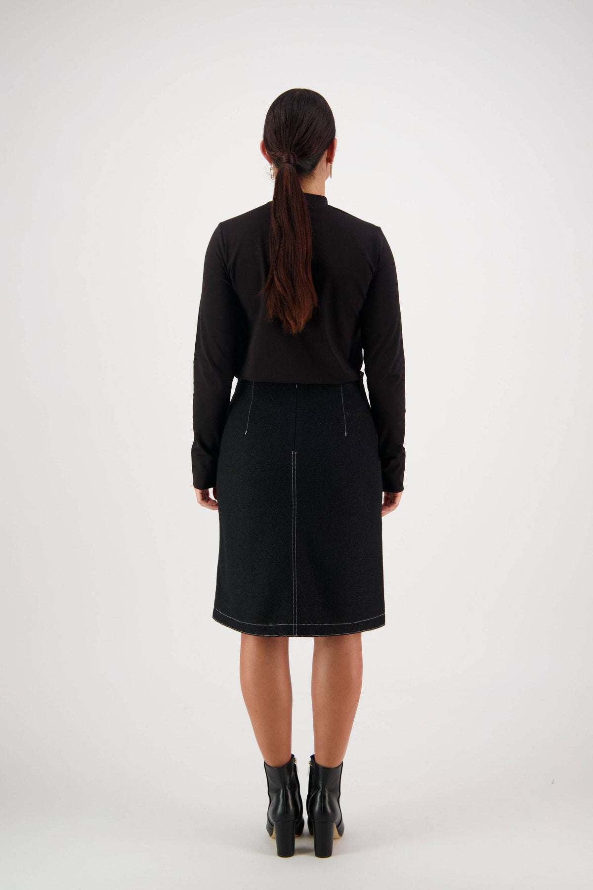 A woman with long brown hair tied in a ponytail stands facing away from the camera, dressed in a black long-sleeve top and the Vassalli Knee Length Skirt – Black, which features white stitching and stylish front patch pockets. She finishes her outfit with black ankle boots.