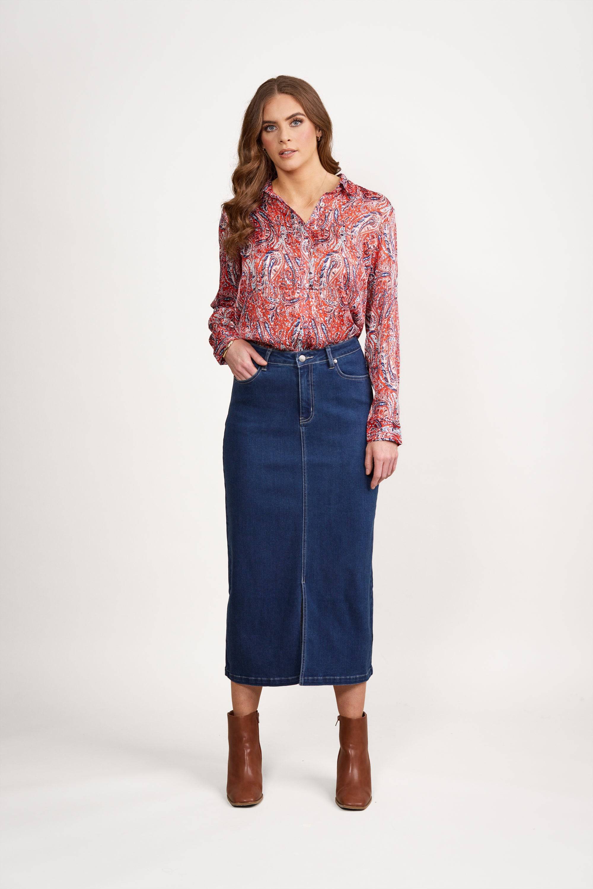 A woman with long, wavy brown hair stands against a white background in a red paisley blouse, Vassalli Mid Length Skirt - Blue Denim, and brown ankle boots. One hand is tucked into her skirt pocket.