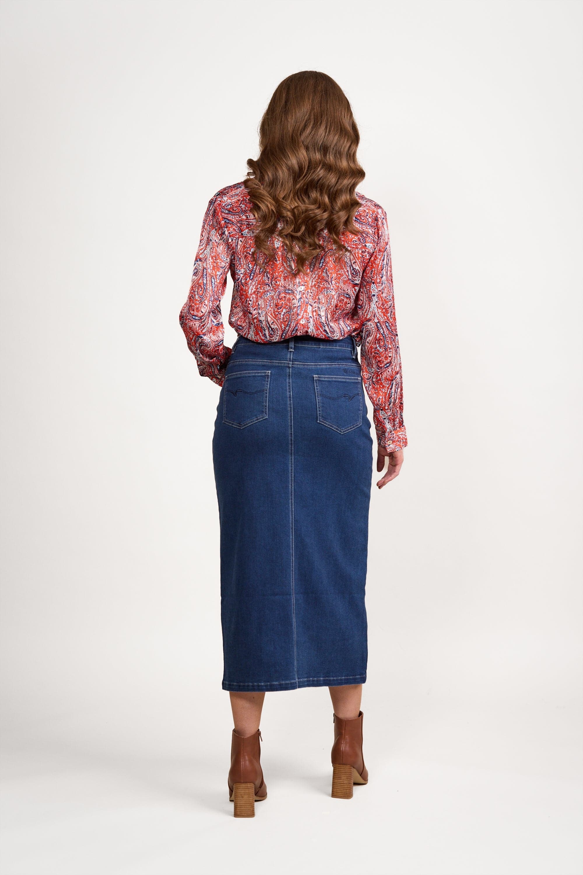 A woman with long wavy hair and a red patterned blouse wears the Vassalli Mid Length Skirt in blue denim and brown heeled ankle boots, standing with her back to the camera against a plain white background.