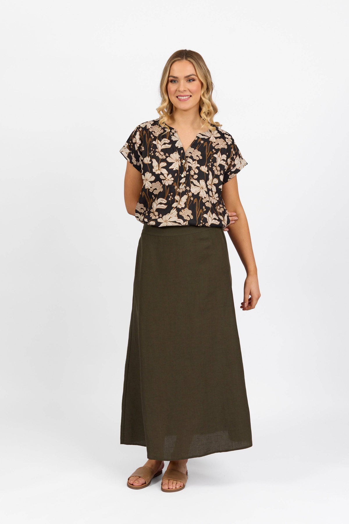 A woman with loose blonde waves smiles against a white background. She wears a black floral blouse, the Vassalli Long Skirt in khaki with an elastic waistband, and tan sandals.