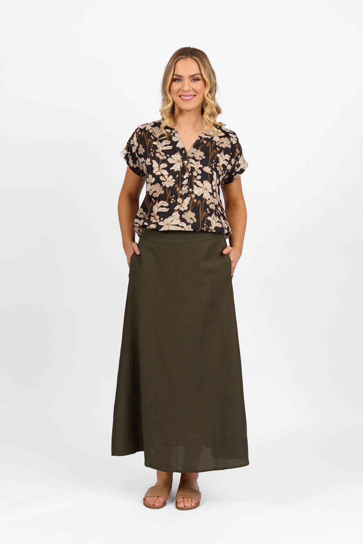 A woman with shoulder-length blonde hair smiles with hands in pockets, wearing a dark floral short-sleeve blouse and a Vassalli Long Skirt in khaki. She wears sandals and stands against a plain white background.