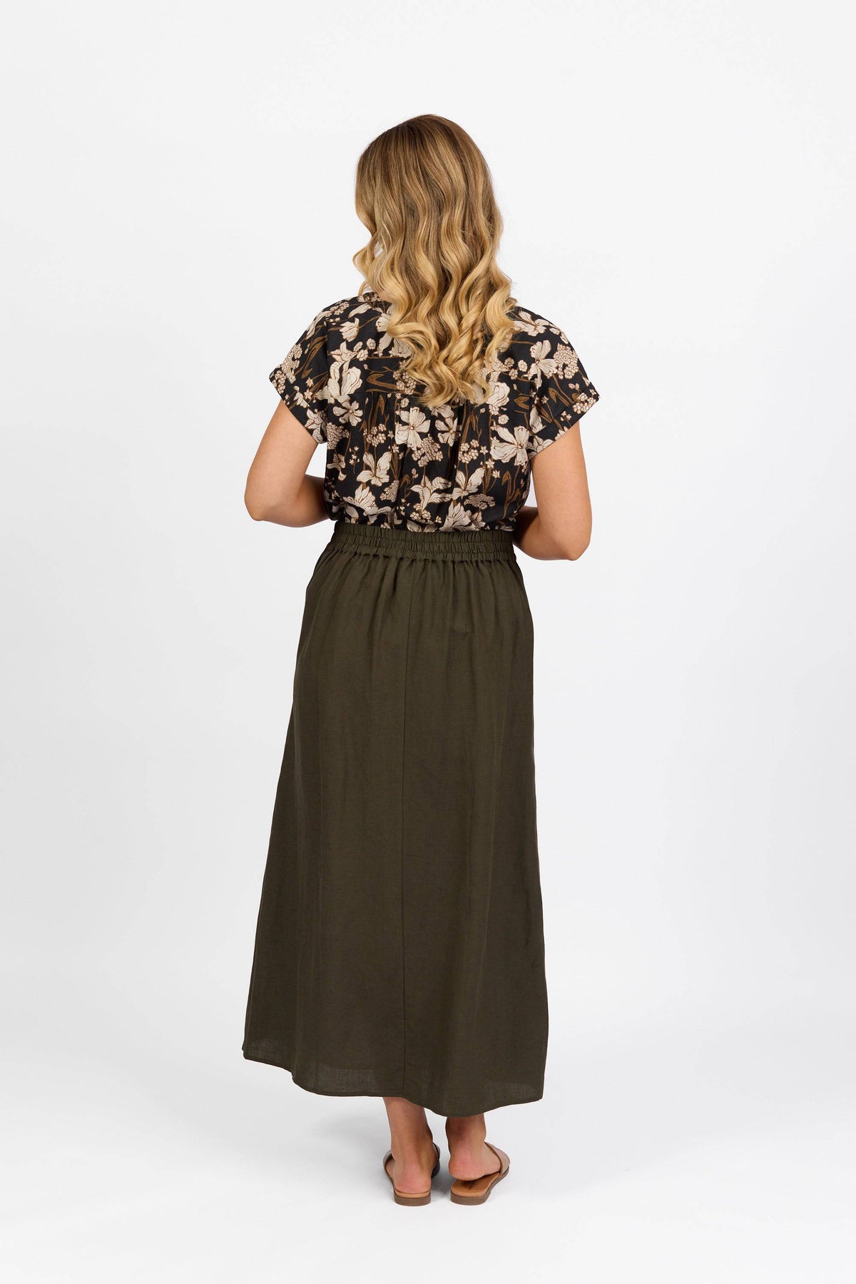 A woman with long, wavy blonde hair stands facing away against a white background, wearing the Vassalli Long Skirt in khaki, paired with a black floral blouse and brown sandals.