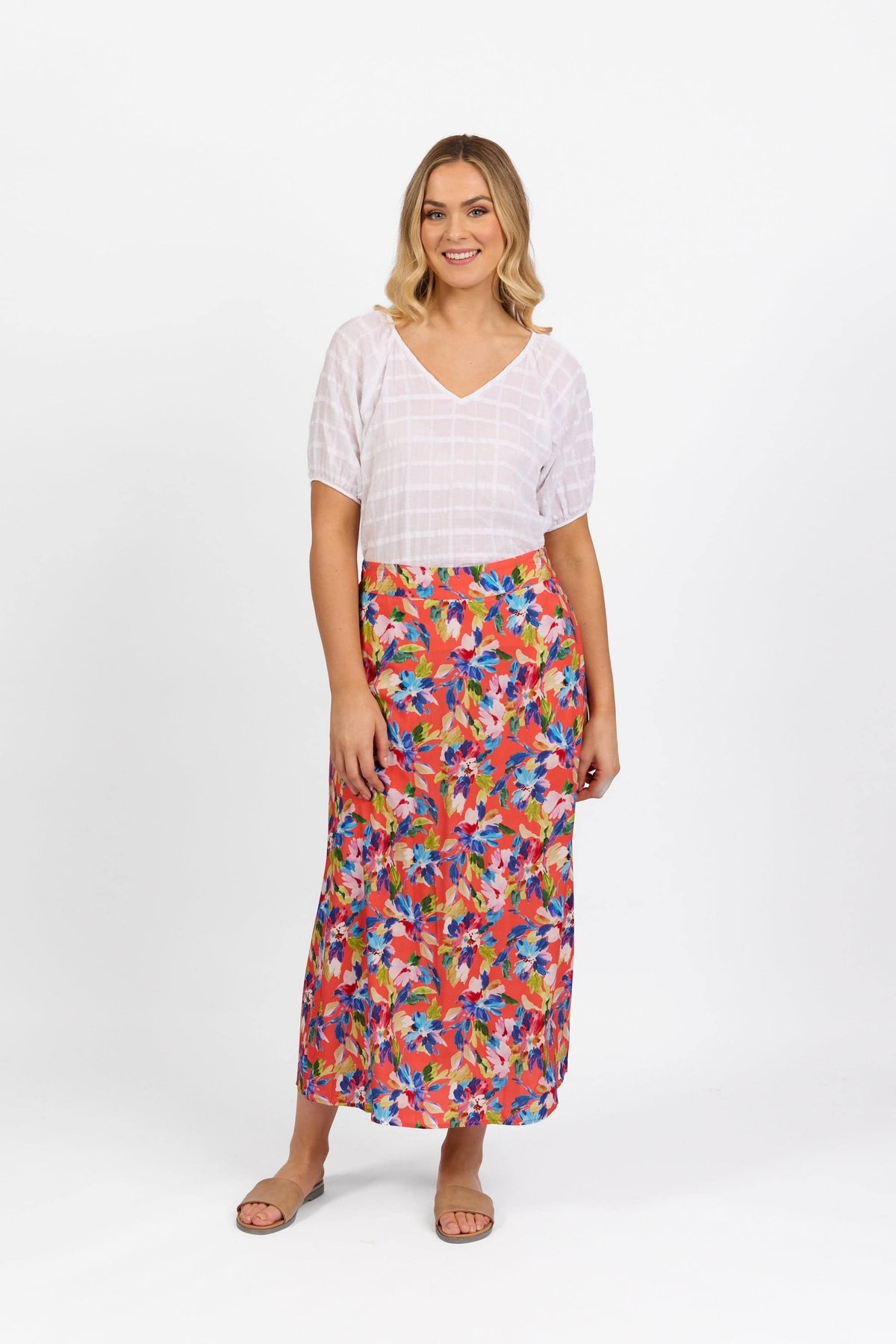 A woman with light hair smiles in a white short-sleeve V-neck top and the Vassalli Long Skirt - Rio, a colorful floral pull-on skirt with an elastic back waistband. She wears beige flat sandals against a plain white background.