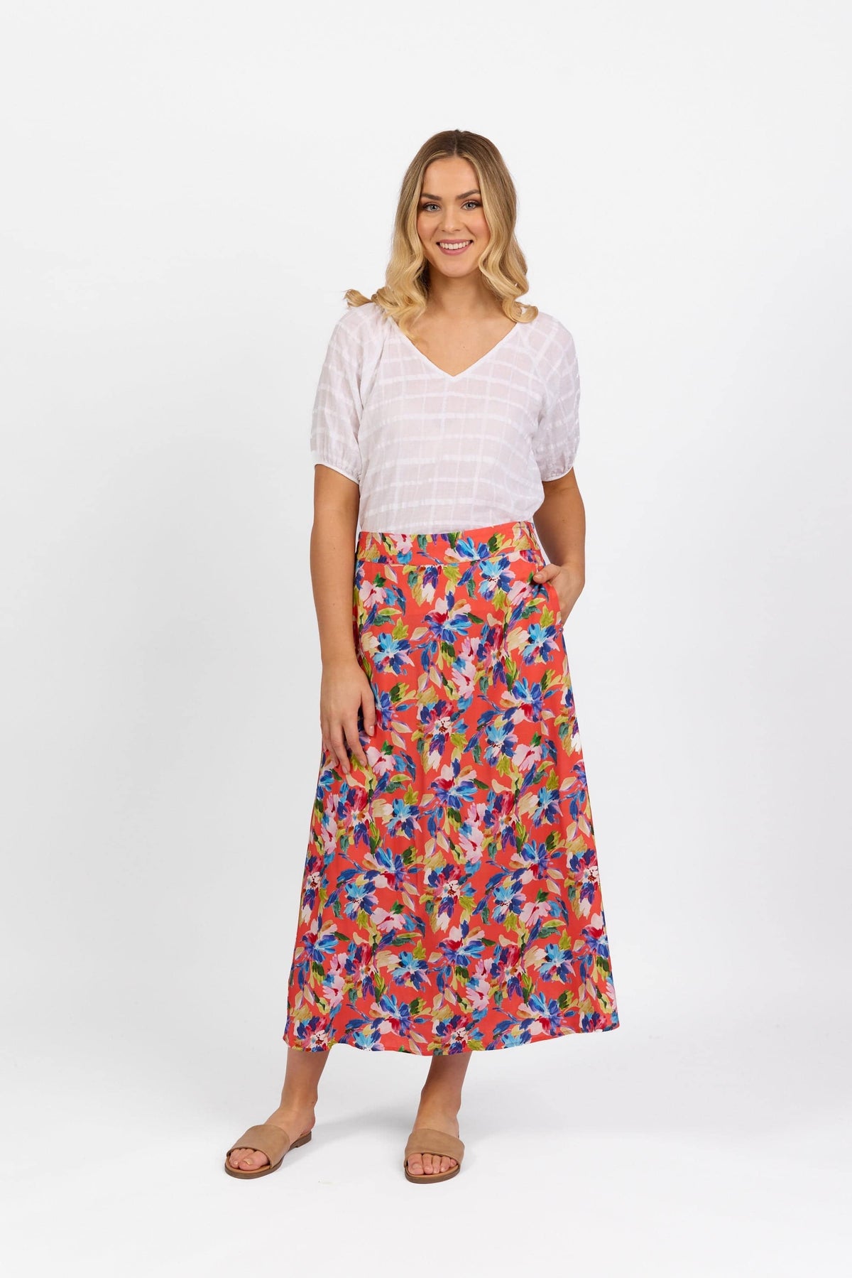 A woman with loose blonde waves smiles, wearing a Vassalli Rio Long Skirt featuring a colorful print, elastic back waistband, and pockets. She pairs it with a white short-sleeve blouse and sandals against a plain white background.