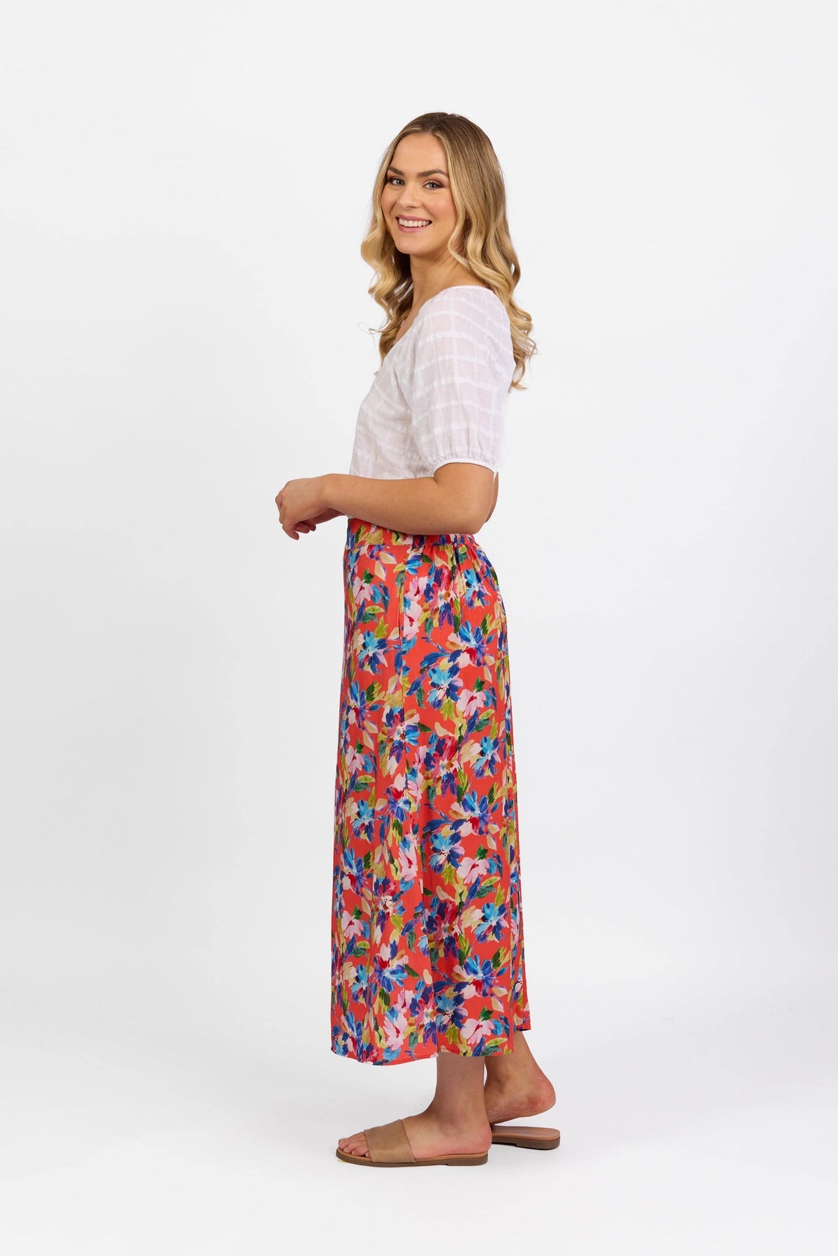 A woman stands sideways, smiling, wearing a white short-sleeve top and the Vassalli Long Skirt - Rio with a colorful floral pattern and elastic back waistband, paired with tan sandals, against a plain white background.