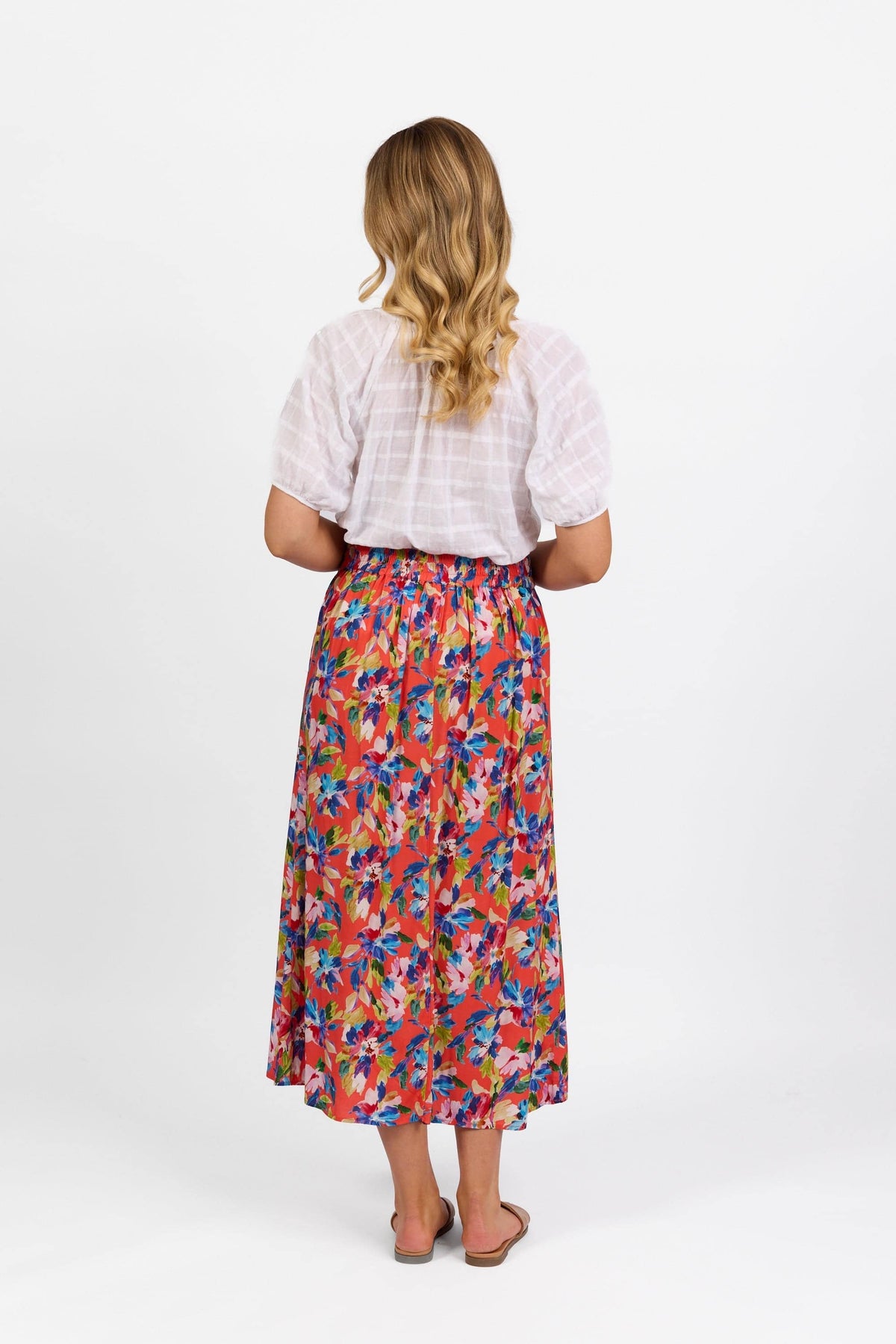 A woman with wavy blonde hair faces away, wearing the Vassalli Long Skirt - Rio, a colorful pull-on style with an elastic back waistband, paired with a white short-sleeve blouse and flat sandals against a plain white background.