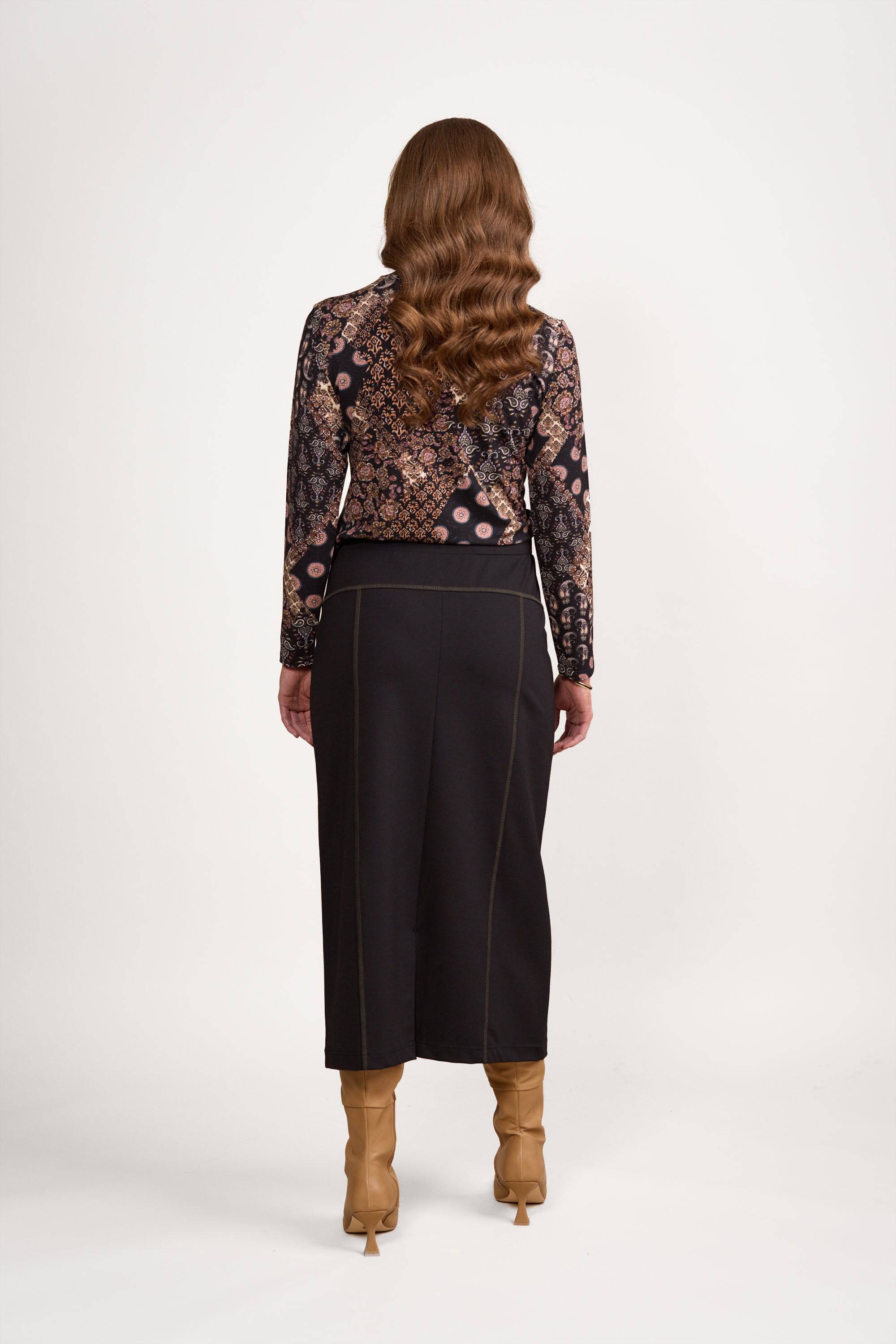 Back view of a woman in a black and gold paisley print long-sleeve top, Vassalli Skirt - Black/Khaki, and tan knee-high heeled boots, shown against a white background.