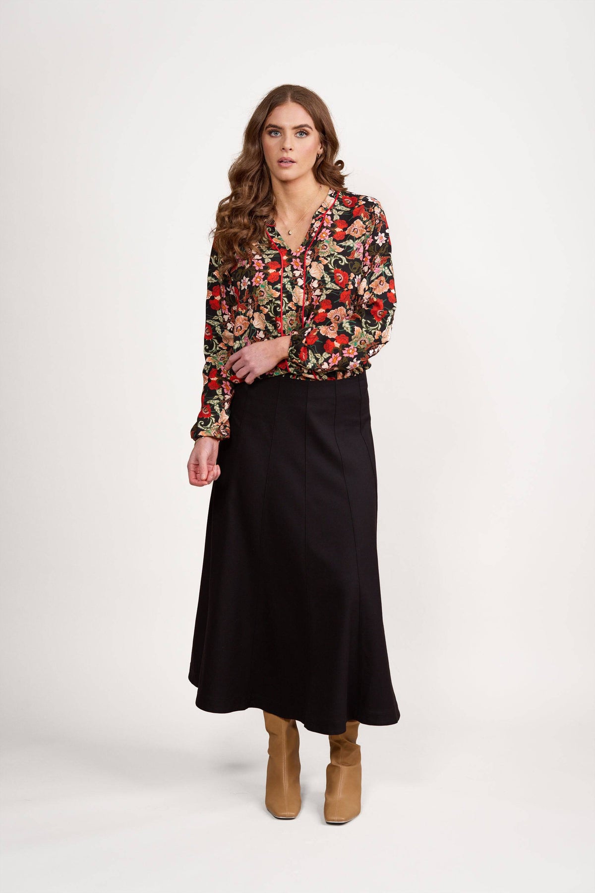 A woman with long, wavy brown hair and a neutral expression stands against a white background wearing the Vassalli Panelled Skirt - Black, paired with a floral blouse in red, green, and cream tones, and tan ankle boots.
