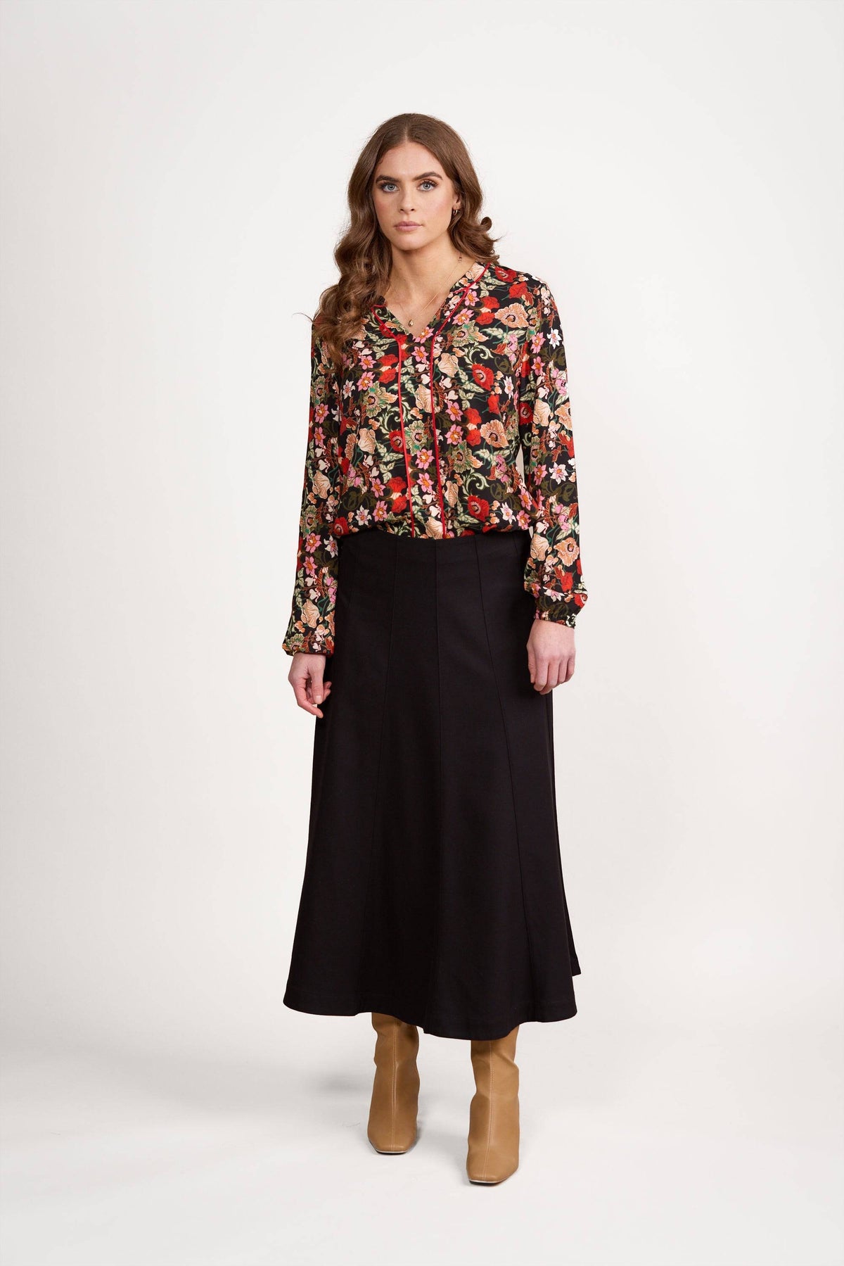 A woman with long wavy hair and a neutral expression stands against a plain white background, wearing the Vassalli Panelled Skirt in black, paired with a floral long-sleeved blouse and tan knee-high boots.