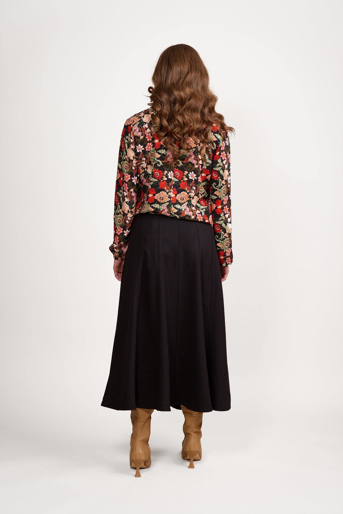 A woman with wavy brown hair stands with her back to the camera, wearing a red floral long-sleeve blouse, tan knee-high boots, and the Vassalli Panelled Skirt in black against a plain white background.
