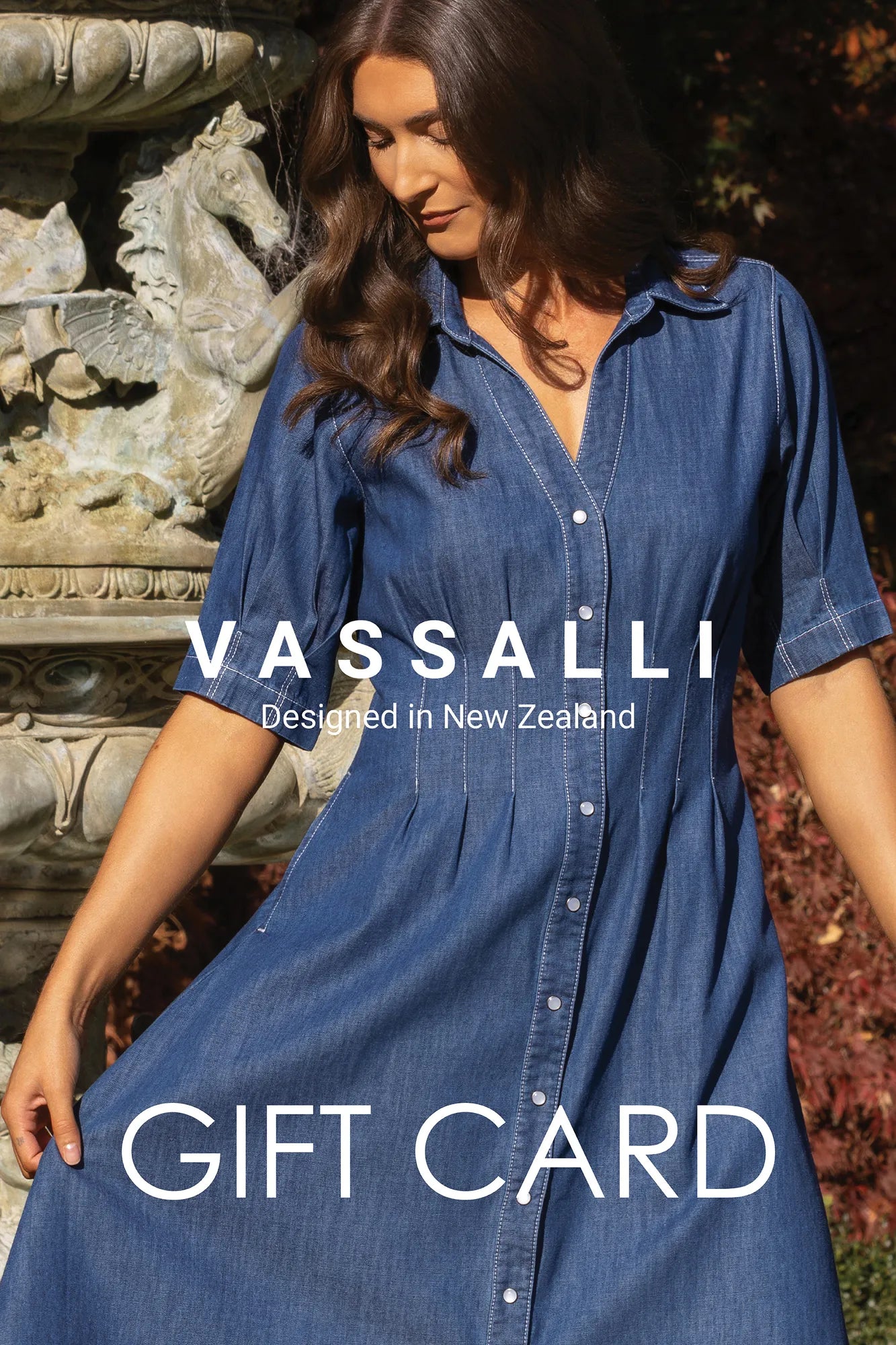 A woman in a blue denim dress stands outdoors near a stone fountain. Text on the image reads: “Vassalli E-Gift Card – designed in New Zealand by Vassalli – making it the perfect gift of choice for any occasion.”.