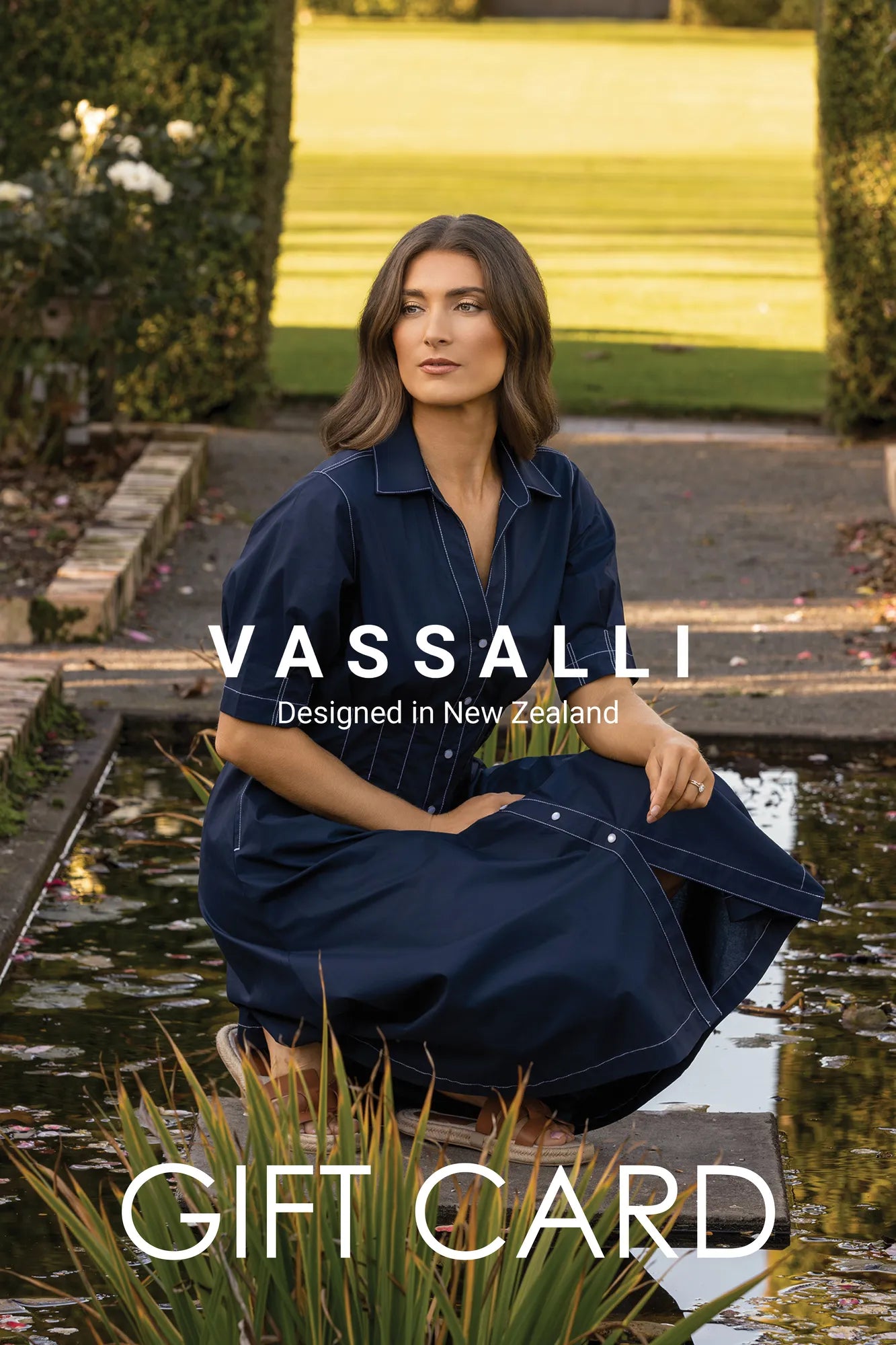 A woman wearing a navy dress sits beside a reflective pond in a lush garden. Text reads: Vassalli E-Gift Card by Vassalli—Designed in New Zealand and the perfect gift of choice.