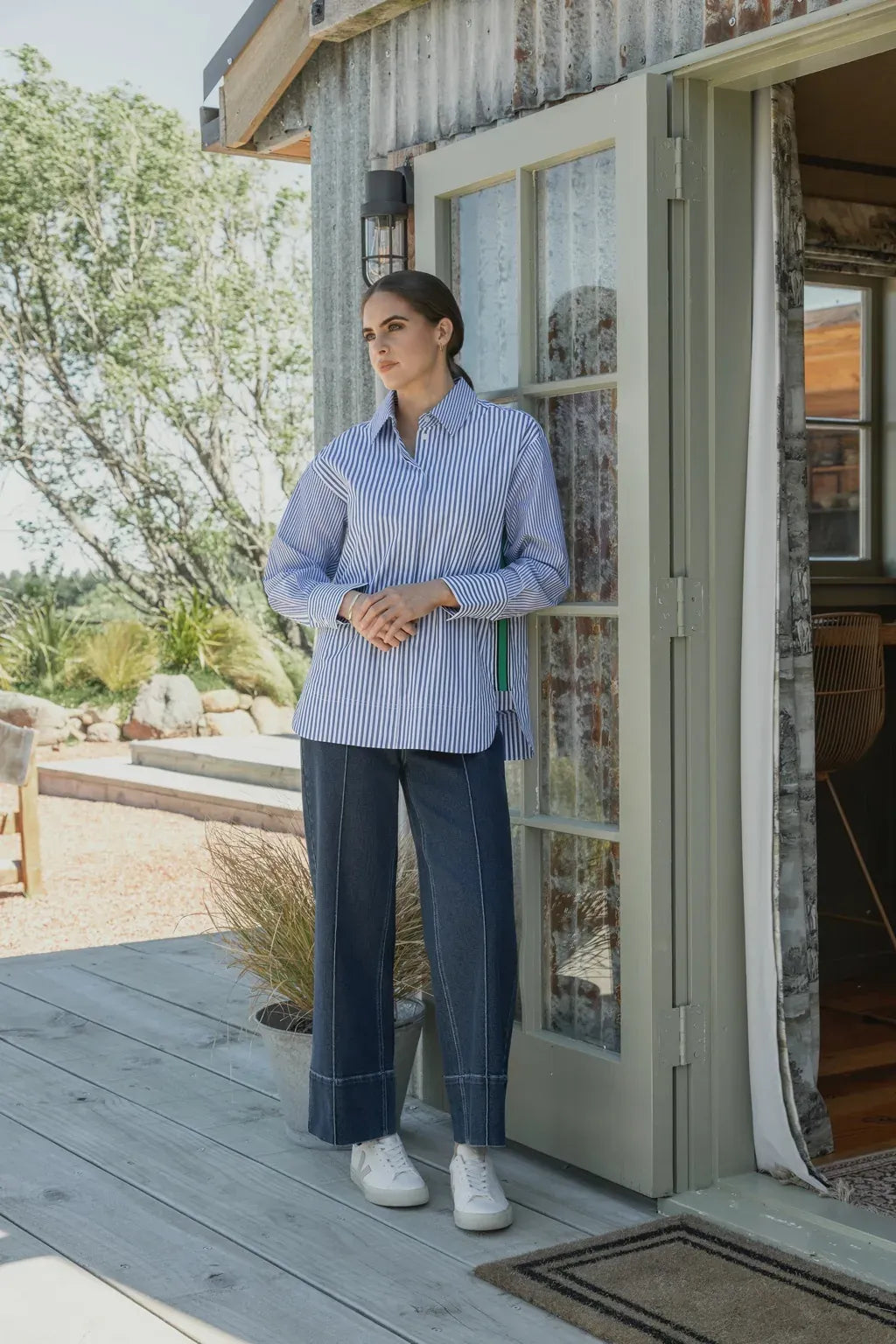 A woman wearing a blue and white striped shirt, dark wide-leg jeans, and white sneakers stands in a doorway, looking outside. She is on a wooden porch with trees and plants in the background.