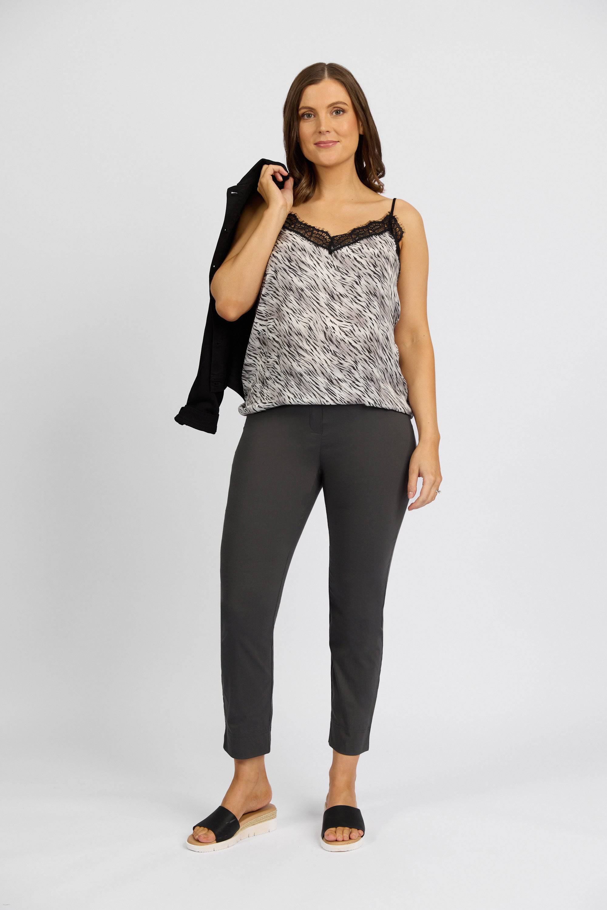 A woman stands against a white background, wearing Vassalli’s Paris Capri Pant in Beluga, paired with a black and white sleeveless top and black slide sandals. She holds a black jacket over her shoulder and smiles slightly.