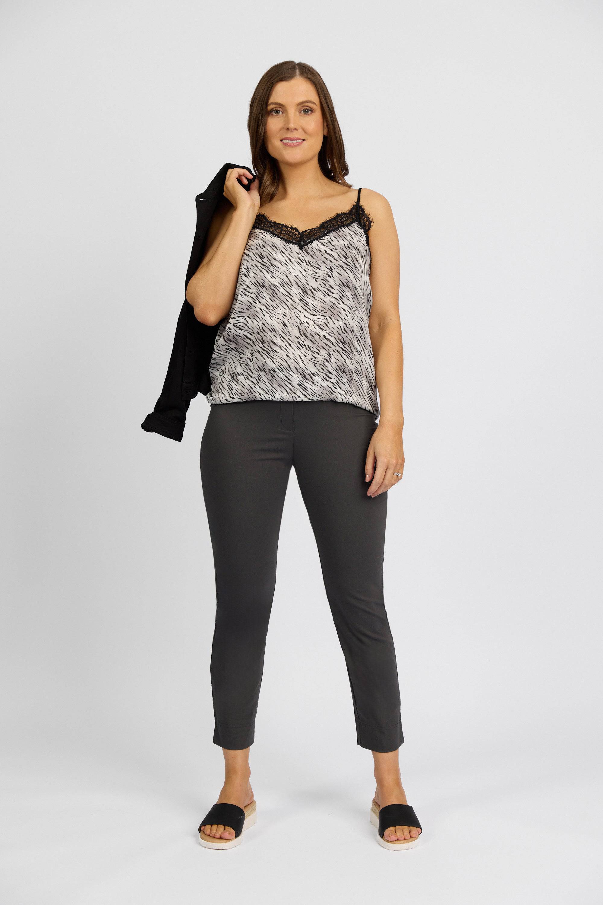 A woman stands against a plain background, wearing a black and white sleeveless top, dark pants, and black sandals. She holds a black jacket over her right shoulder and smiles slightly—her stretch capri pants are the Paris Capri Pant - Beluga by Vassalli.