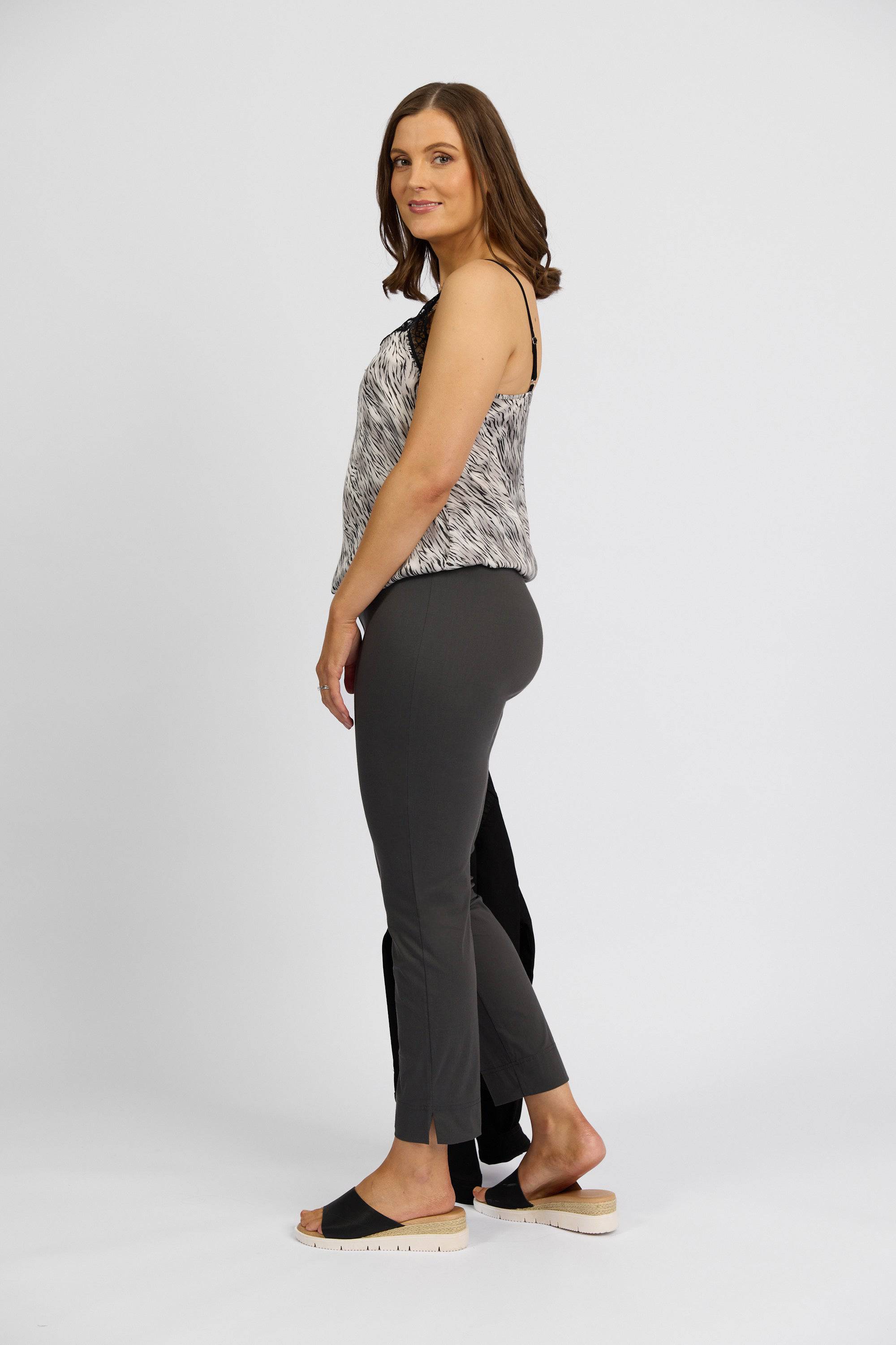 A woman with shoulder-length brown hair stands sideways, smiling softly in Vassalli's Paris Capri Pant - Beluga, paired with a sleeveless patterned top and light sandals, channeling effortless Paris Collection style against a plain background.