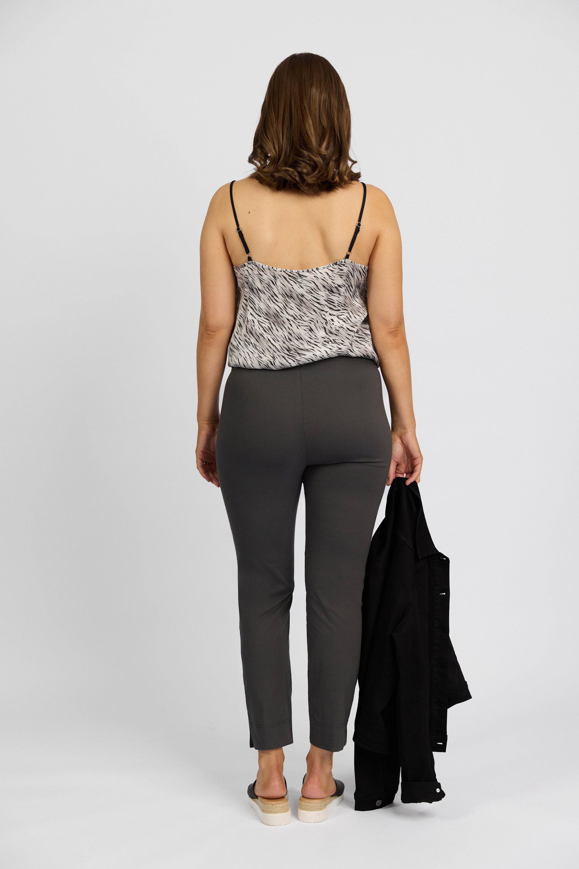 A woman with shoulder-length brown hair faces away, wearing a black and white sleeveless top, Vassalli Paris Capri Pant - Beluga, light shoes, and holding a black jacket in her left hand against a plain white background.