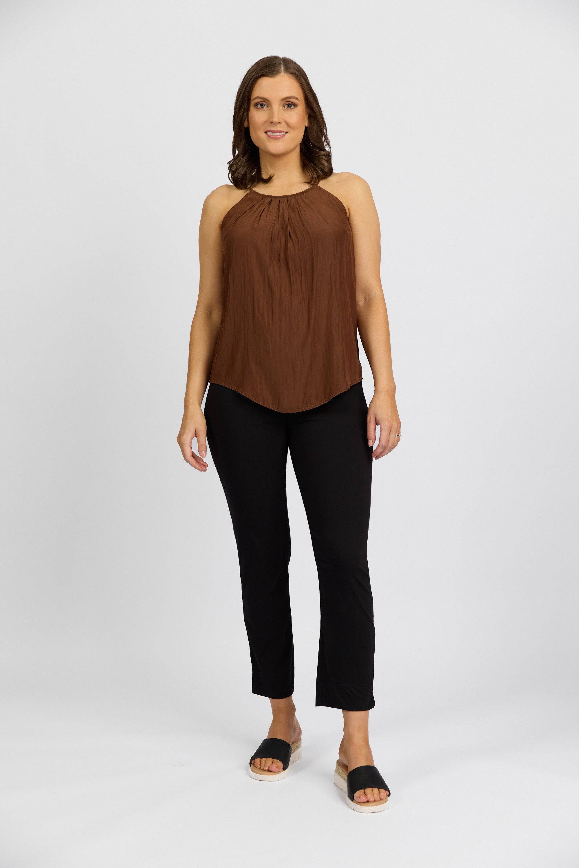 A woman stands against a white background wearing a brown sleeveless top and Vassalli Paris Capri Pant - Black, featuring a mid rise 7/8 fit in stretch fabric, paired with black slide sandals. She smiles with her arms relaxed at her sides.