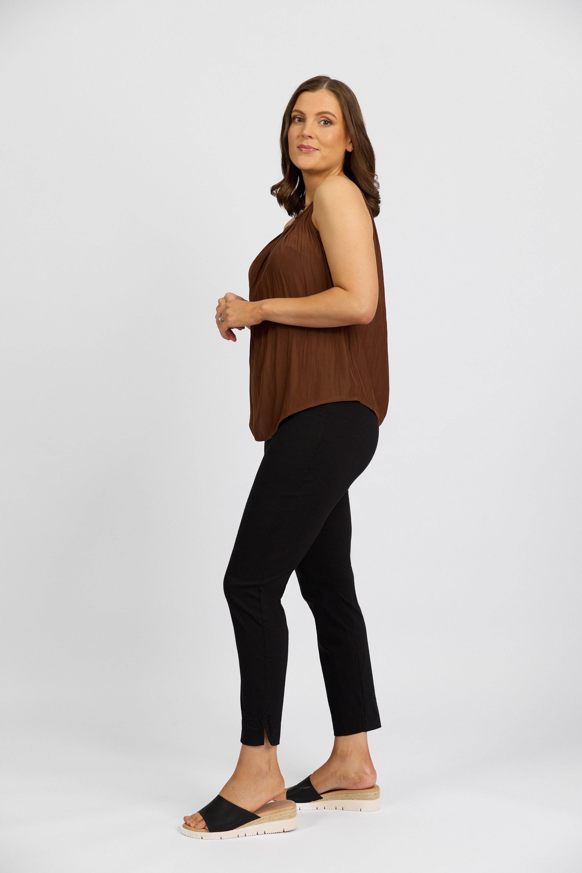 A woman with shoulder-length brown hair stands sideways, wearing a sleeveless brown top and Vassalli's Paris Capri Pant - Black in a mid-rise 7/8 stretch fit, paired with black slide sandals with white soles against a white background.